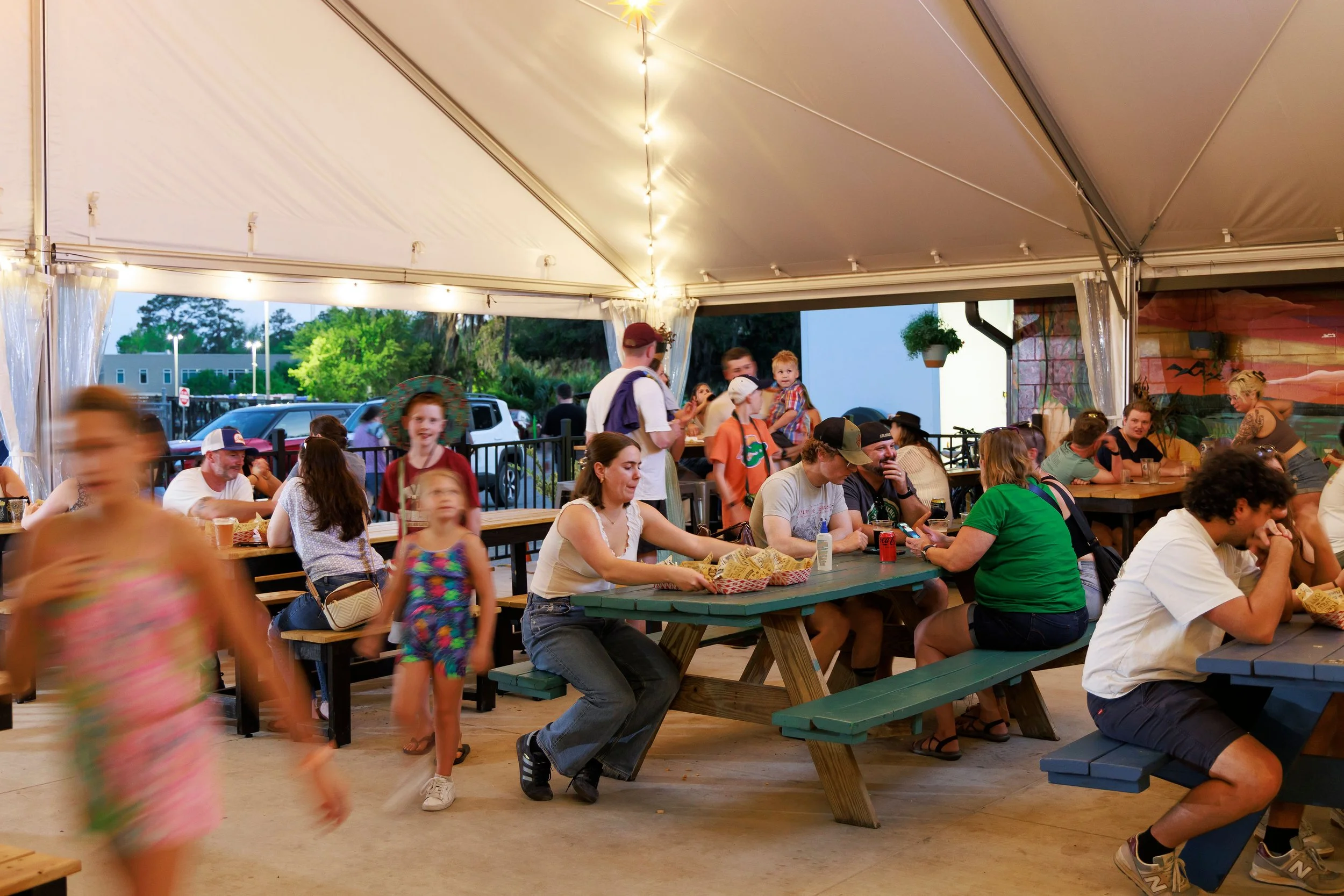 People attending a gathering inside a tent, sitting at picnic tables, with some standing and walking. The tent has string lights and is open at the sides showing a parking lot and trees outside.