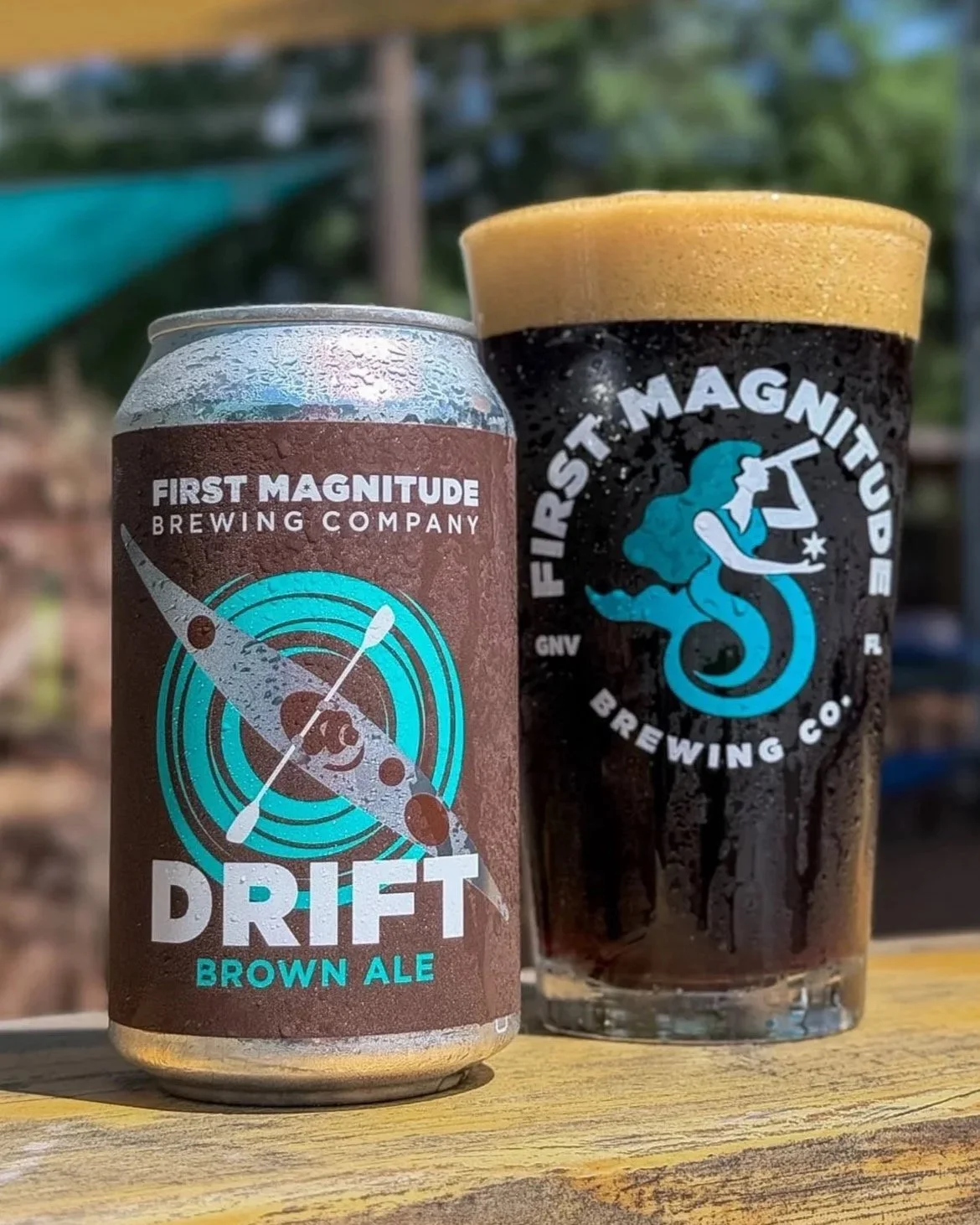 A can of First Magnitude Brewing Company's Drift Brown Ale and a glass of dark beer with the First Magnitude Brewing Co. logo on it, with a blurred outdoor background.