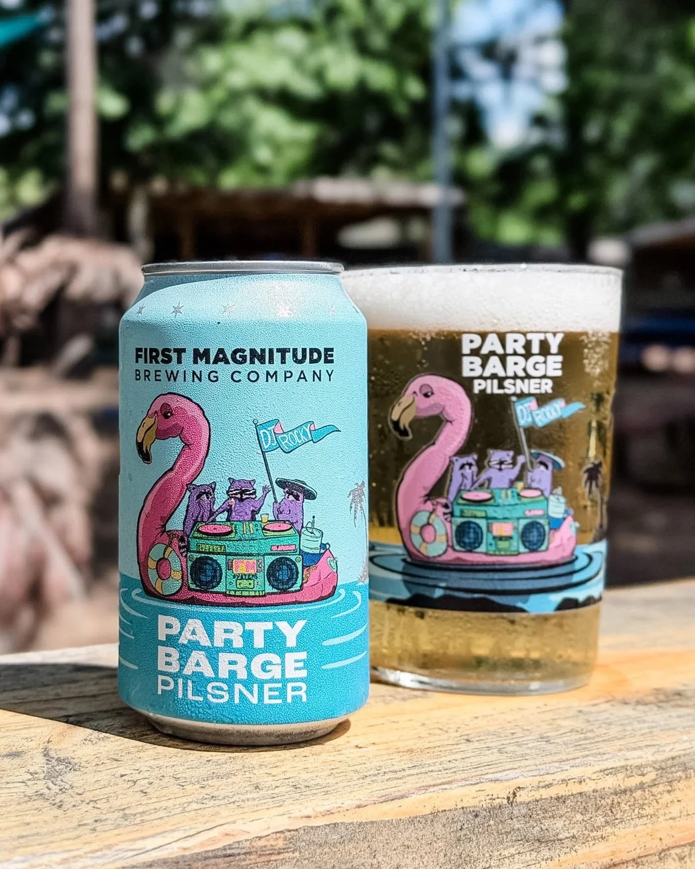 A can of Party Barge Pilsner beer and a glass of beer on a wooden surface outdoors, with a background of trees and green foliage.