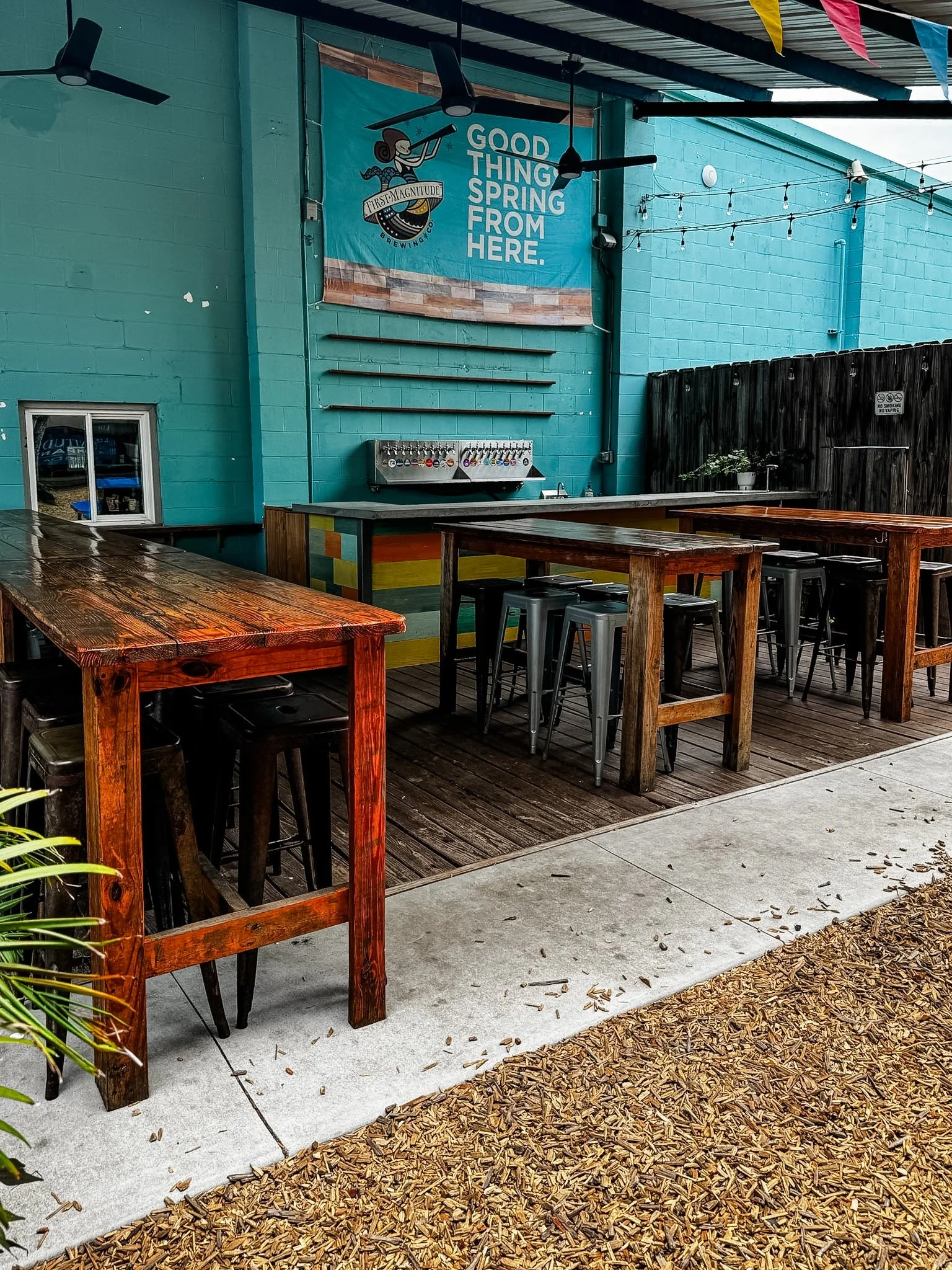 Outdoor patio area with wooden tables, metal stools, string lights, a teal wall with a brewing company banner, and a gravel and concrete ground.