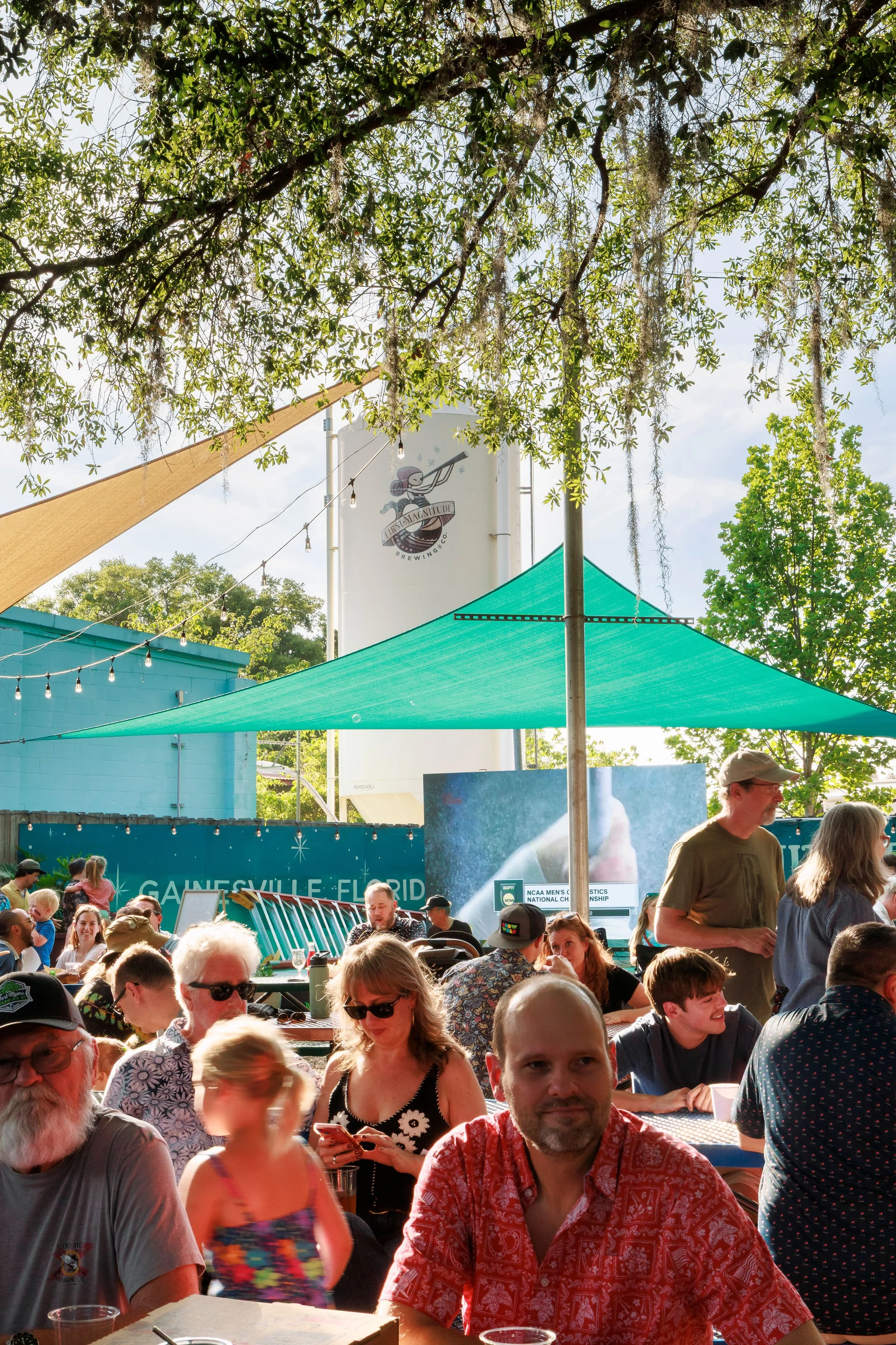 Outdoor event with a crowd of people gathered under a green shade, some seated and some standing. There are string lights hanging, and a banner that says "Gainesville Florida." Trees and a building are visible in the background, with a sign for First Magnitude Brewing Co.