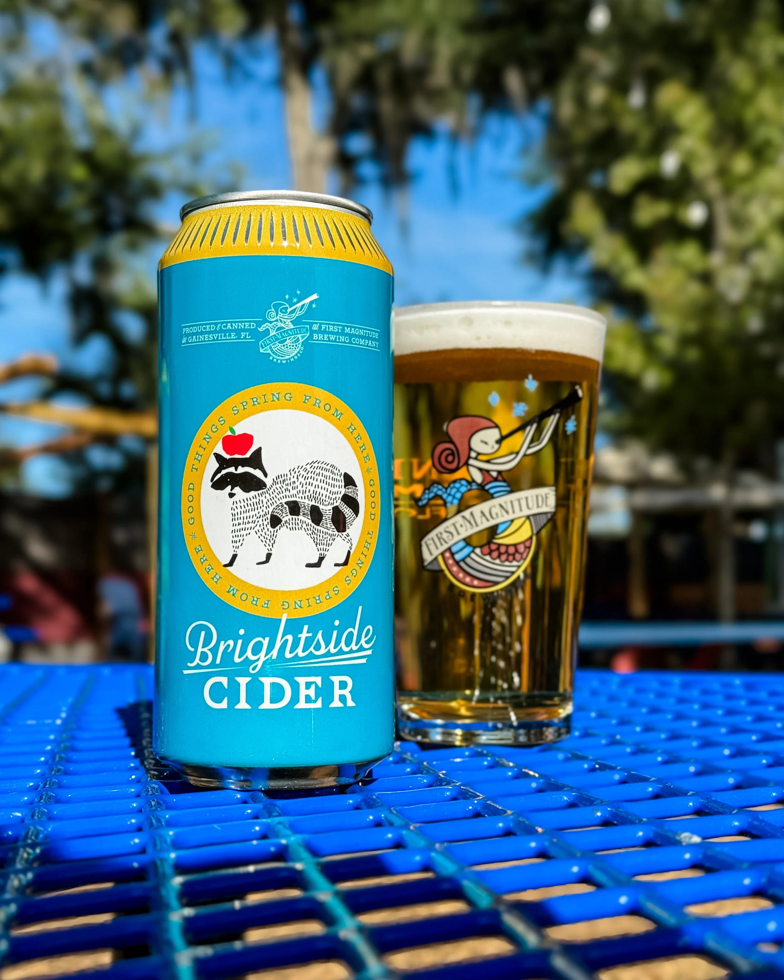 A can of Brightside cider and a glass of beer sitting on a blue outdoor table, with trees and blue sky in the background.