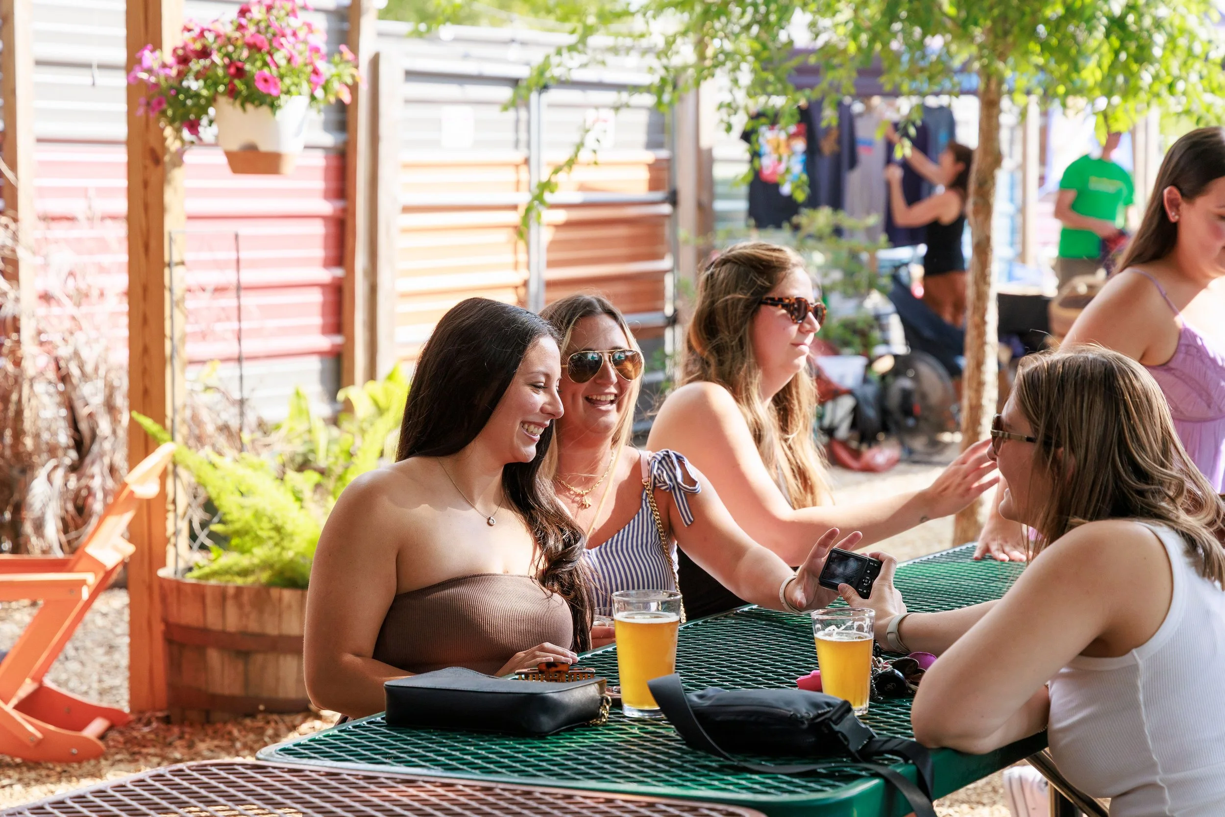 Group of women sitting at a green outdoor table with drinks during daytime, socializing in a sunny backyard patio with plants, trees, and a wooden fence.