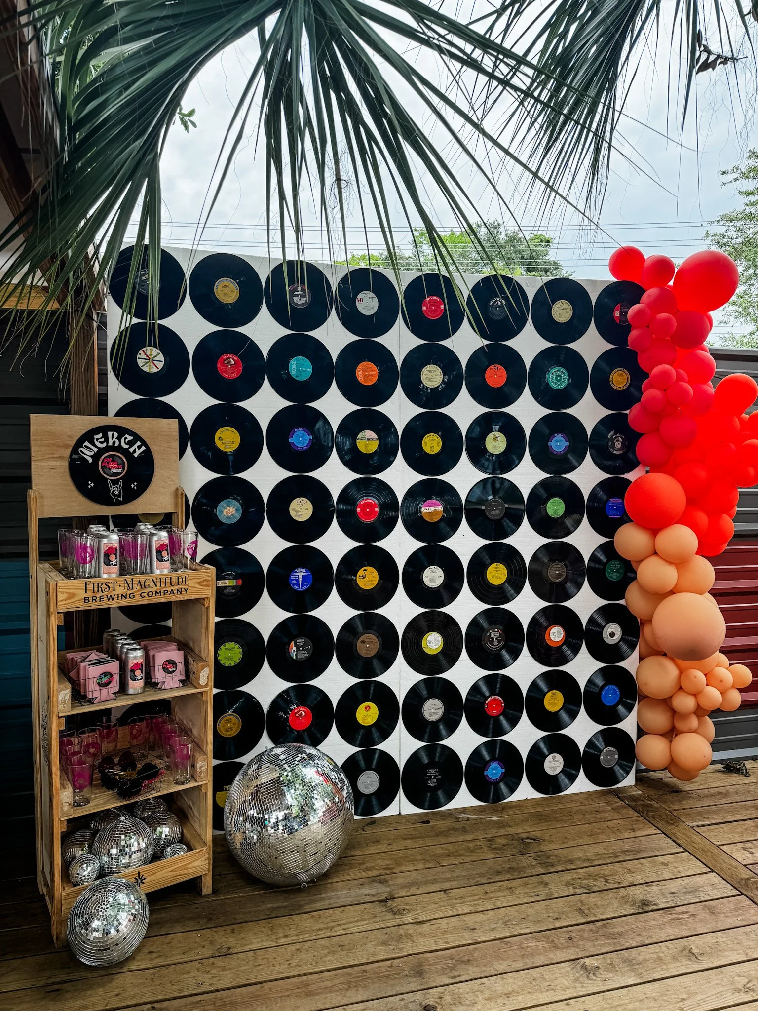 A wall decorated with vinyl records, a cluster of red and peach balloons on the right, a disco ball on the floor, and a wooden shelf with glasses and pink boxes. Part of a palm tree is visible at the top left, and there is a wooden deck floor.