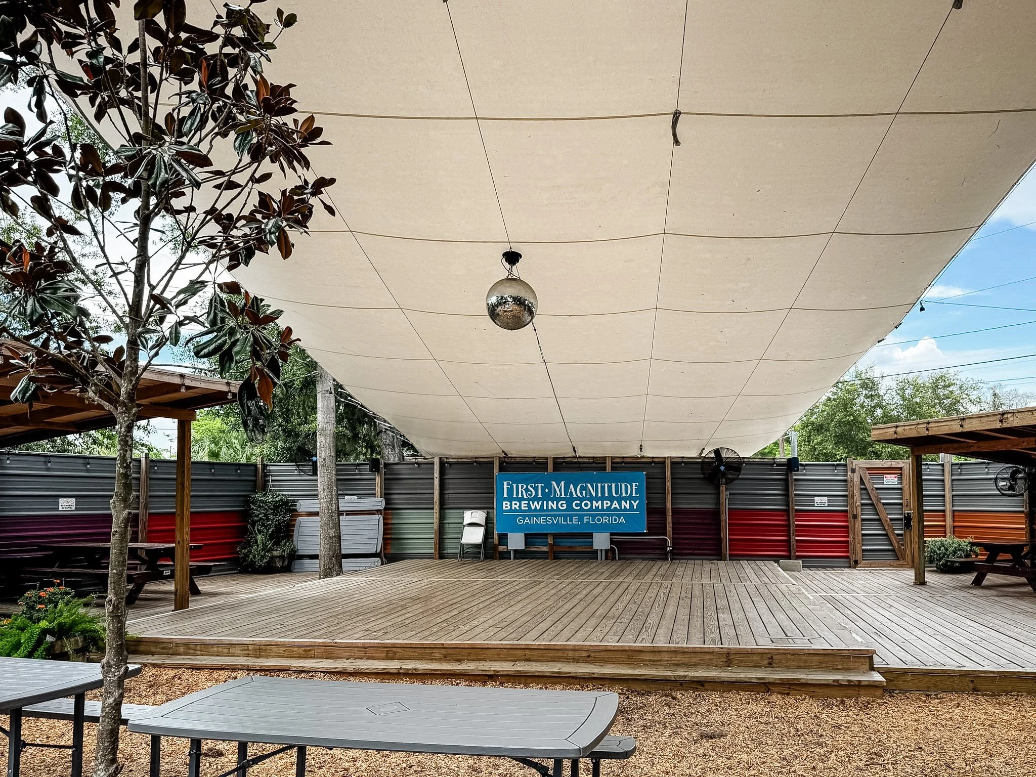 Outdoor area with wooden deck, trees, string lights, and a sign for First Magnitude Brewing Company in Gainesville, Florida. A large fabric canopy covers part of the space, with a disco ball hanging underneath.
