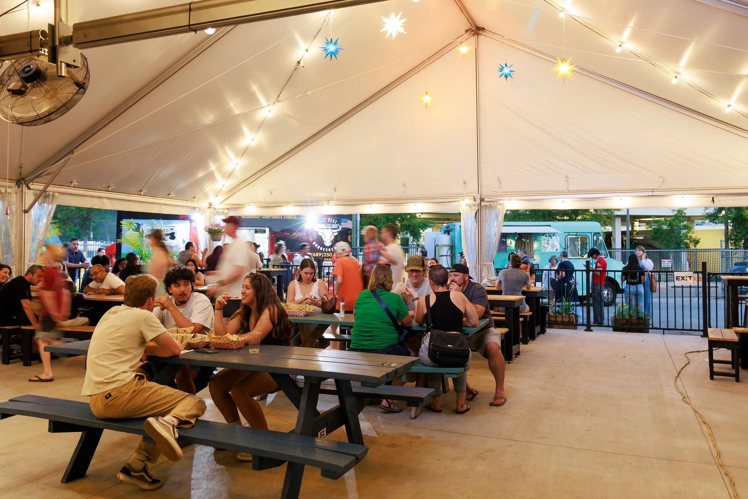 People sitting at picnic tables inside a large tent with string lights and star-shaped decorations, food trucks outside, some individuals walking and socializing.