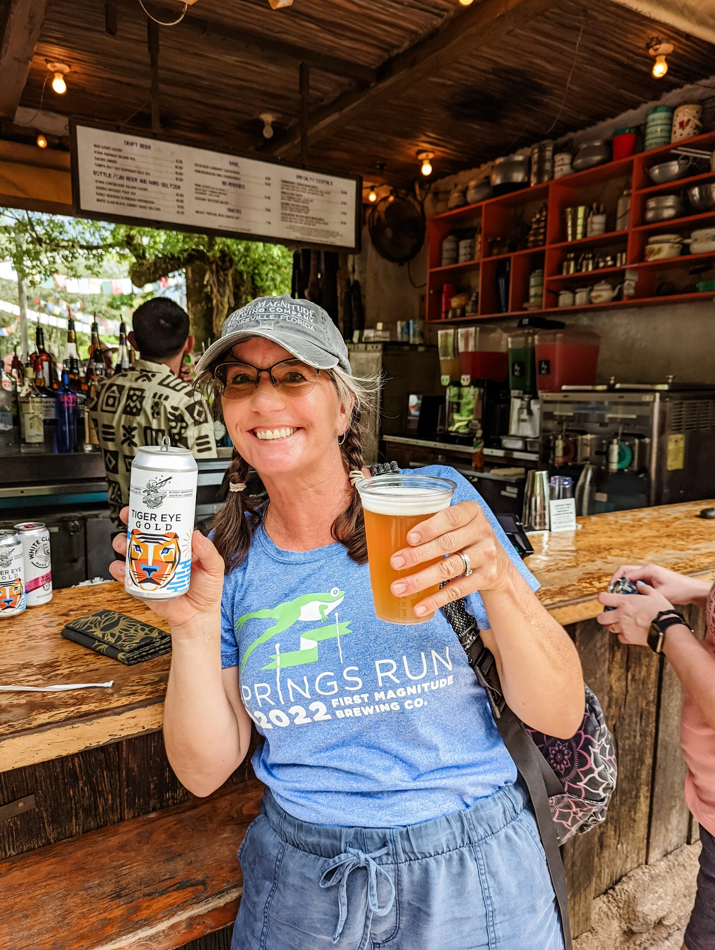 A woman smiling and holding a can of Tiger Eye Gold beer and a glass of beer inside a rustic bar. She is wearing glasses, a gray hat, and a blue t-shirt with a lizard graphic, and blue shorts. Behind her are shelves with plates and bowls, and a large menu hanging on the ceiling. Other people are seen in the background, including a man wearing a patterned shirt.