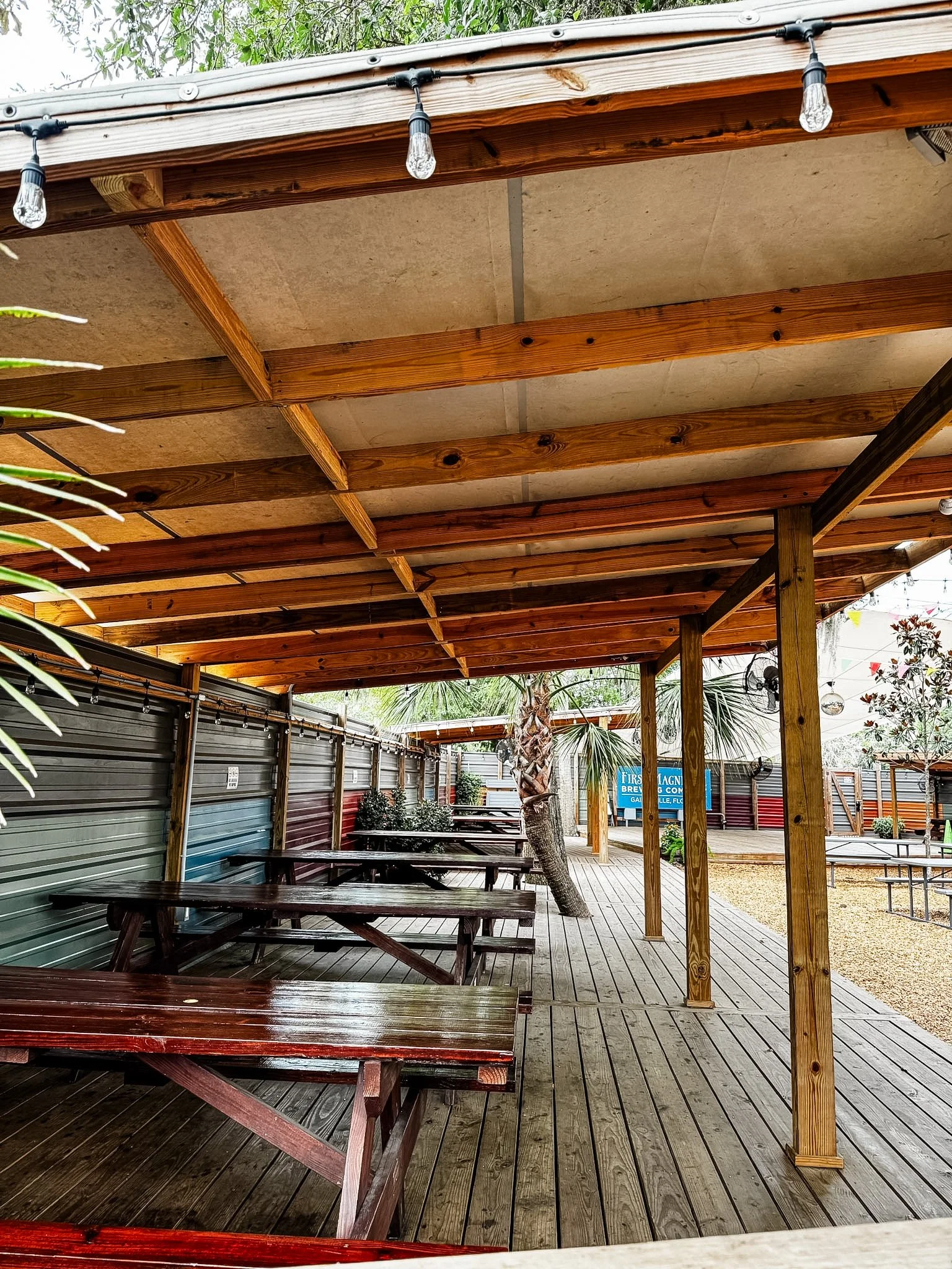 Outdoor patio area with wooden tables and benches, a wooden pergola with string lights, a metal fence, and landscaped plants including palm trees.