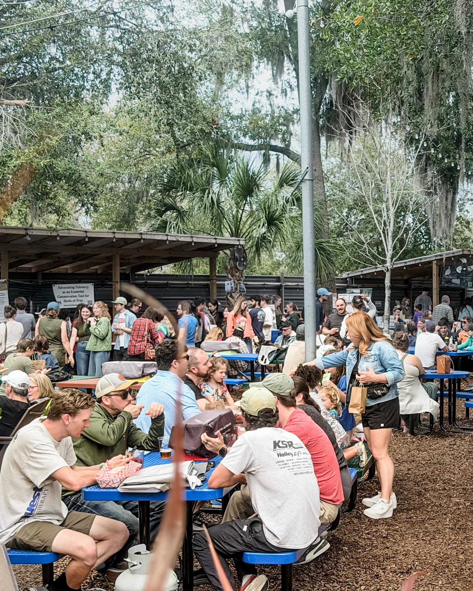 Crowd of people sitting and standing around blue tables outdoors in a wooded area at a social gathering or event, some eating and chatting.