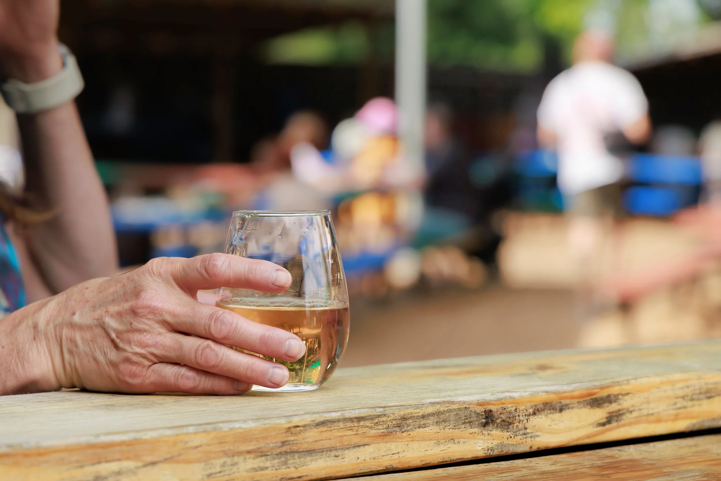 Person holding a wine glass filled with a light-colored beverage, with a blurred outdoor setting and other people in the background.