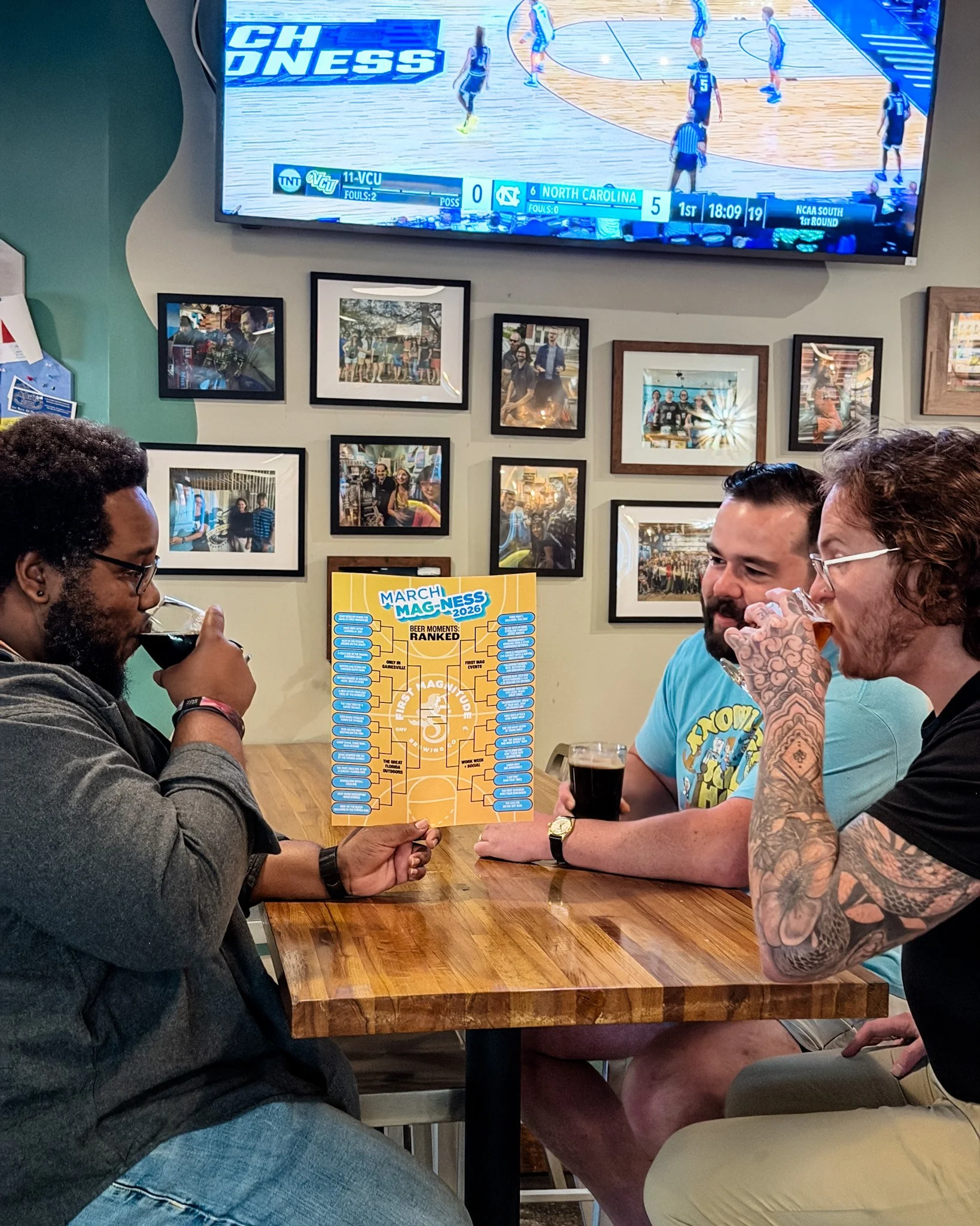 People sitting at a wooden table, holding a March Madness bracket, watching a basketball game on TV in the background.