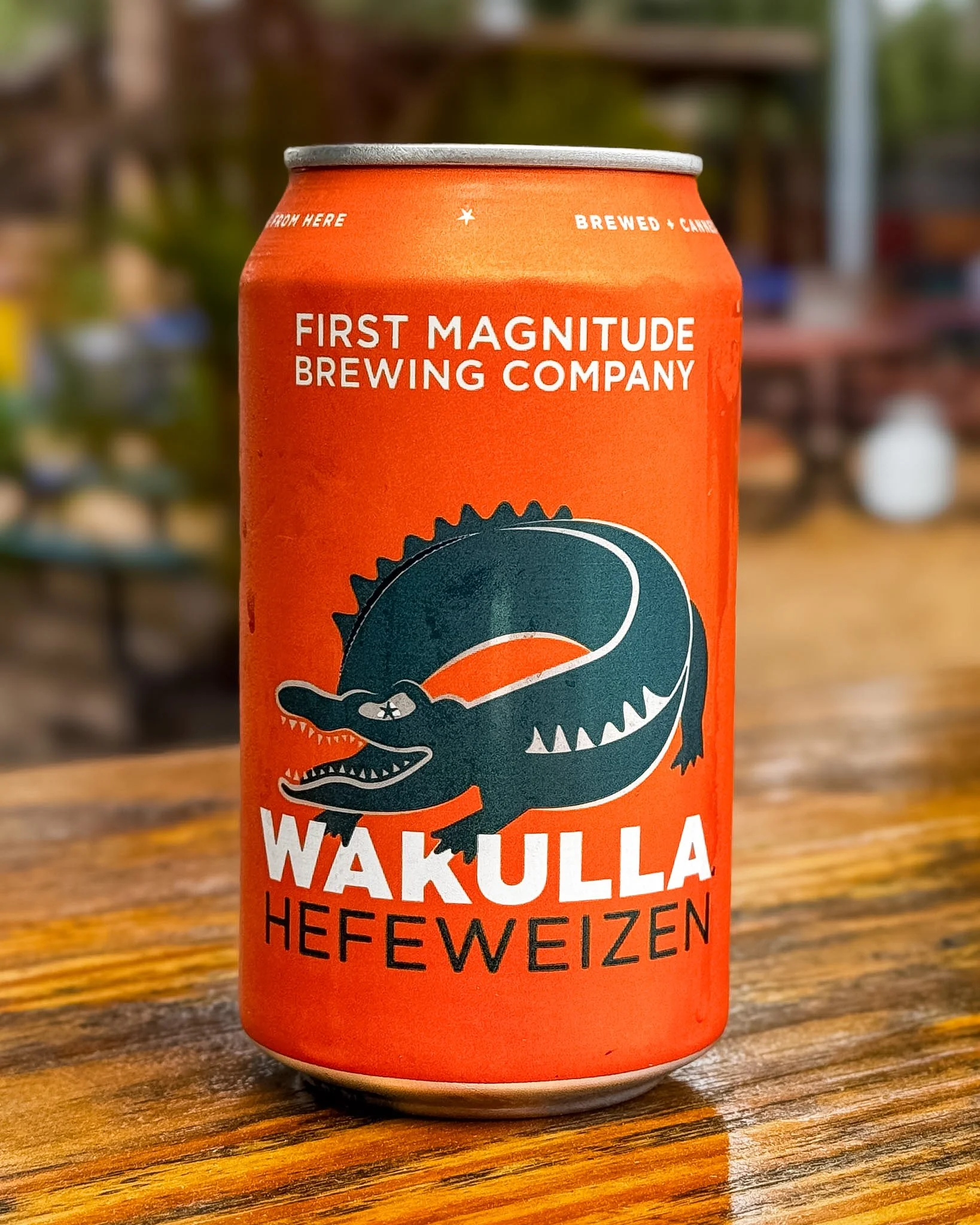 Close-up of a bright orange can of Walker's Hefeweizen beer with a cartoon alligator logo, on a wooden surface outdoors.