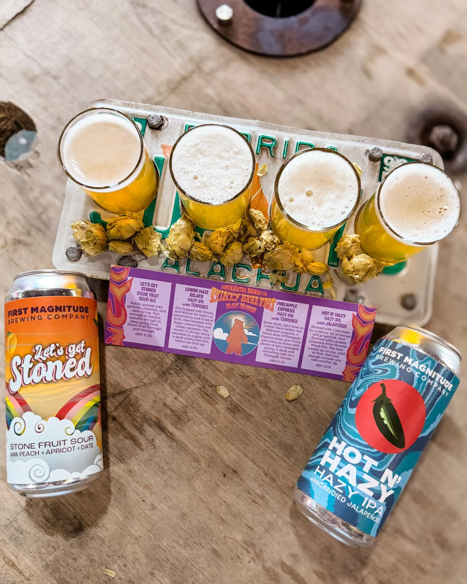 Four glasses of beer, two bottles of flavored beer, and a candy pack on a wooden surface. The beers have foam on top. The bottles are labeled Lemon Haze and Hot'N Hazzy IPA with candied jalapeños.