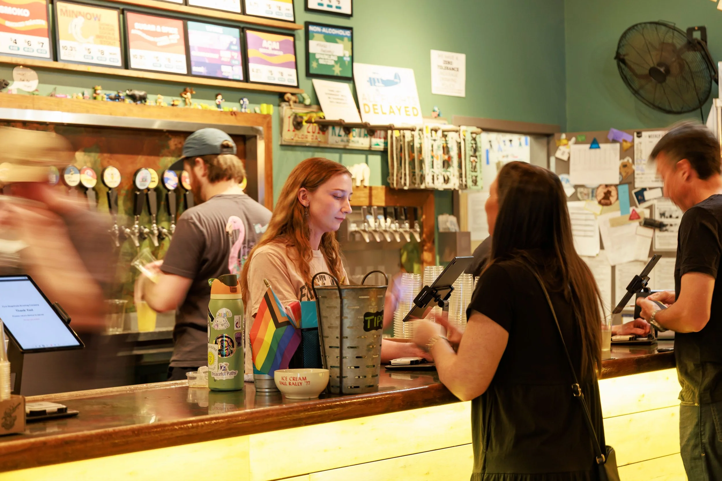 People standing at a counter in a colorful casual restaurant or cafe, with multiple customers being served and a green wall with various signs and menus in the background.