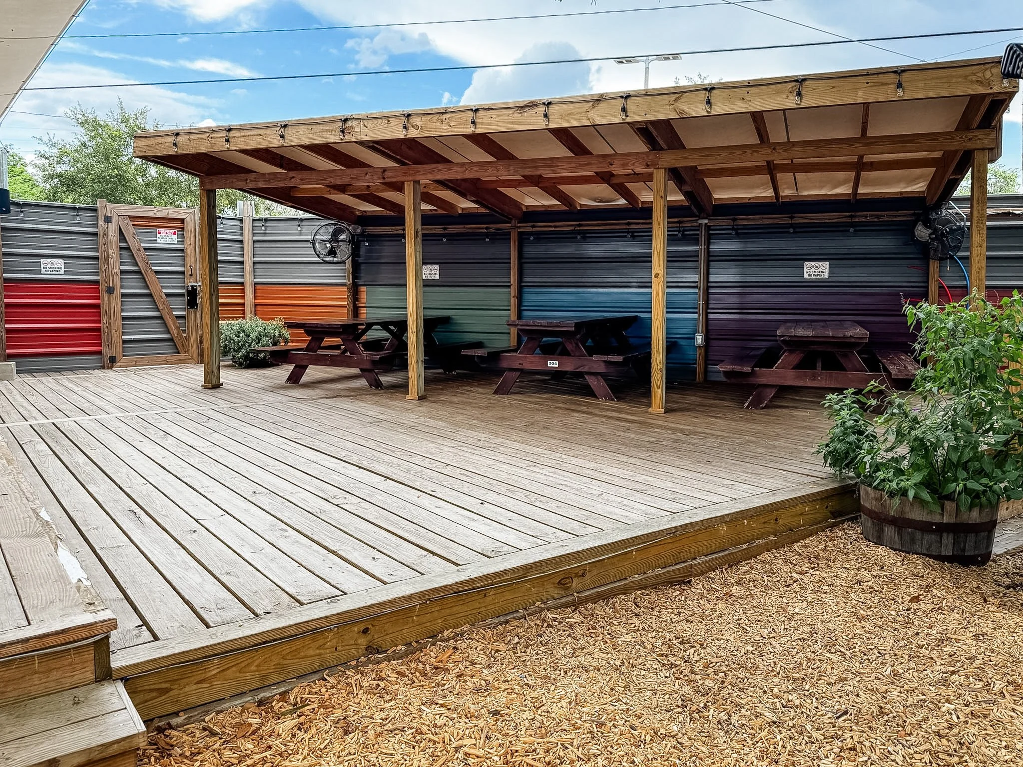 Wooden outdoor patio with a covered section, picnic tables, and decorative string lights against a bright sky with some clouds.