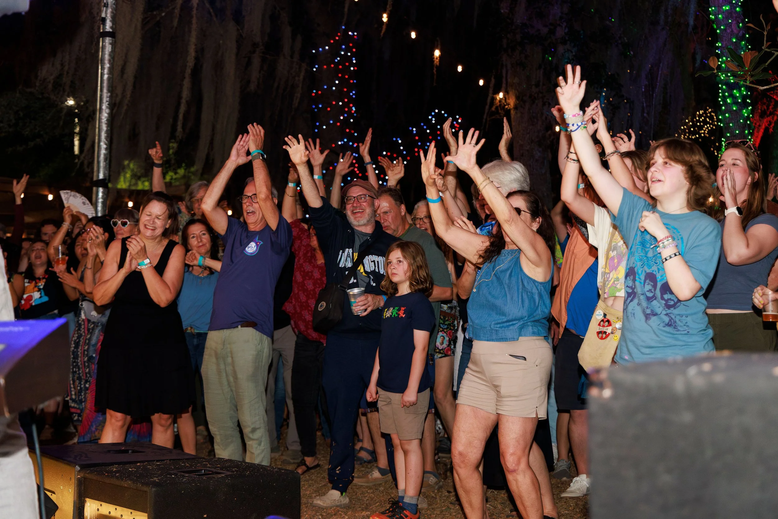 A group of people dancing and enjoying themselves at an outdoor nighttime event with colorful string lights and a decorated tree in the background.