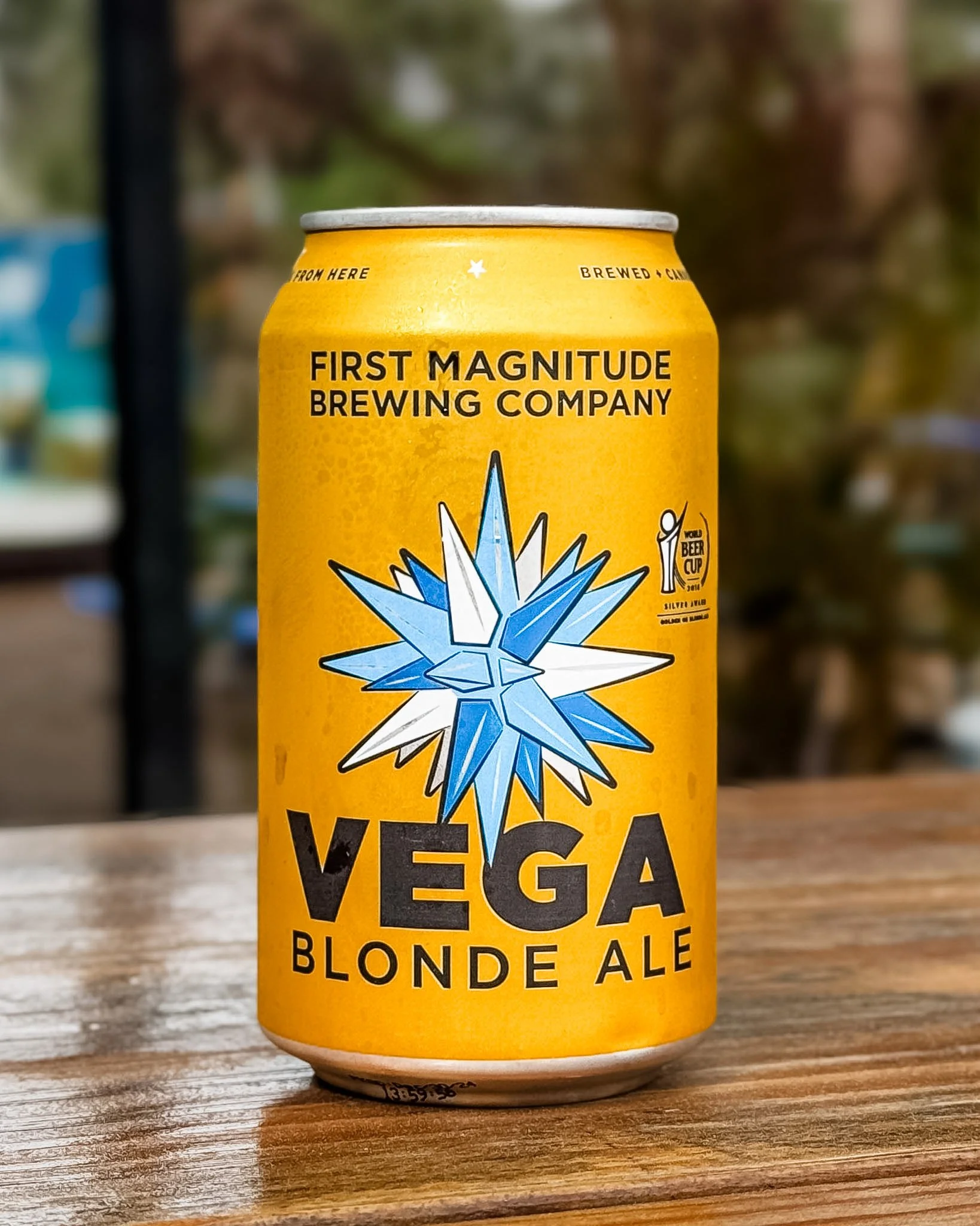 Yellow can of First Magnitude Brewing Company Blonde Ale with a blue and white starburst logo, placed on a wooden surface outdoors with blurred background.