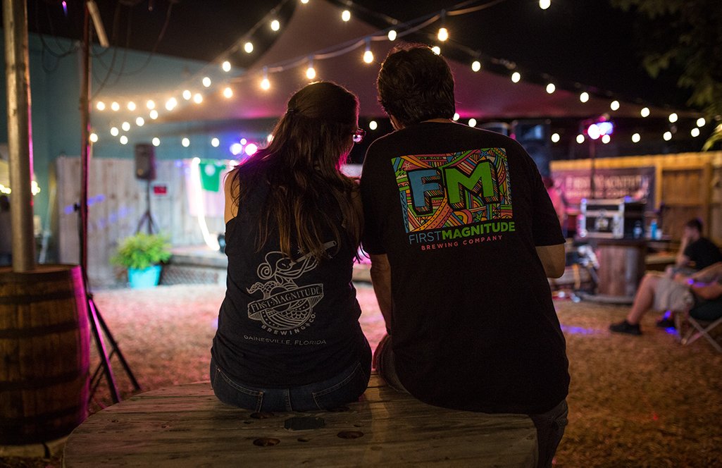 Two people sitting on a wooden bench at night, under string lights, at an outdoor brewery event.
