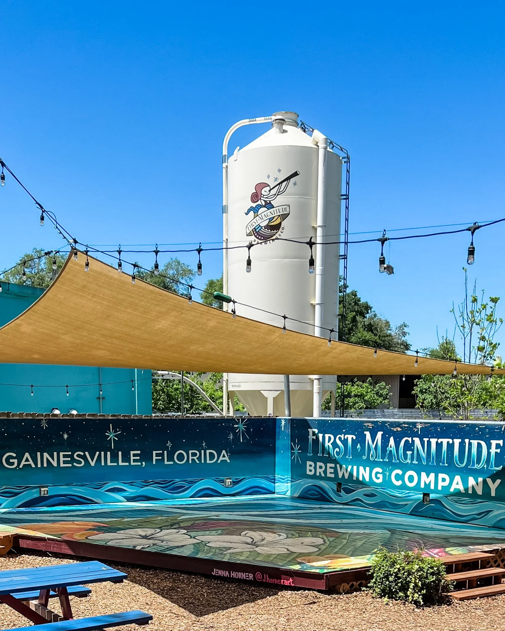 Outdoor scene with a large vertical white water tower featuring a cartoon mermaid looking through a telescope, with the words "First Magnitude Brewing Company" and "Gainesville, Florida" visible. In the foreground, there are string lights, a tan canopy, wooden benches, and a decorated ground surface.