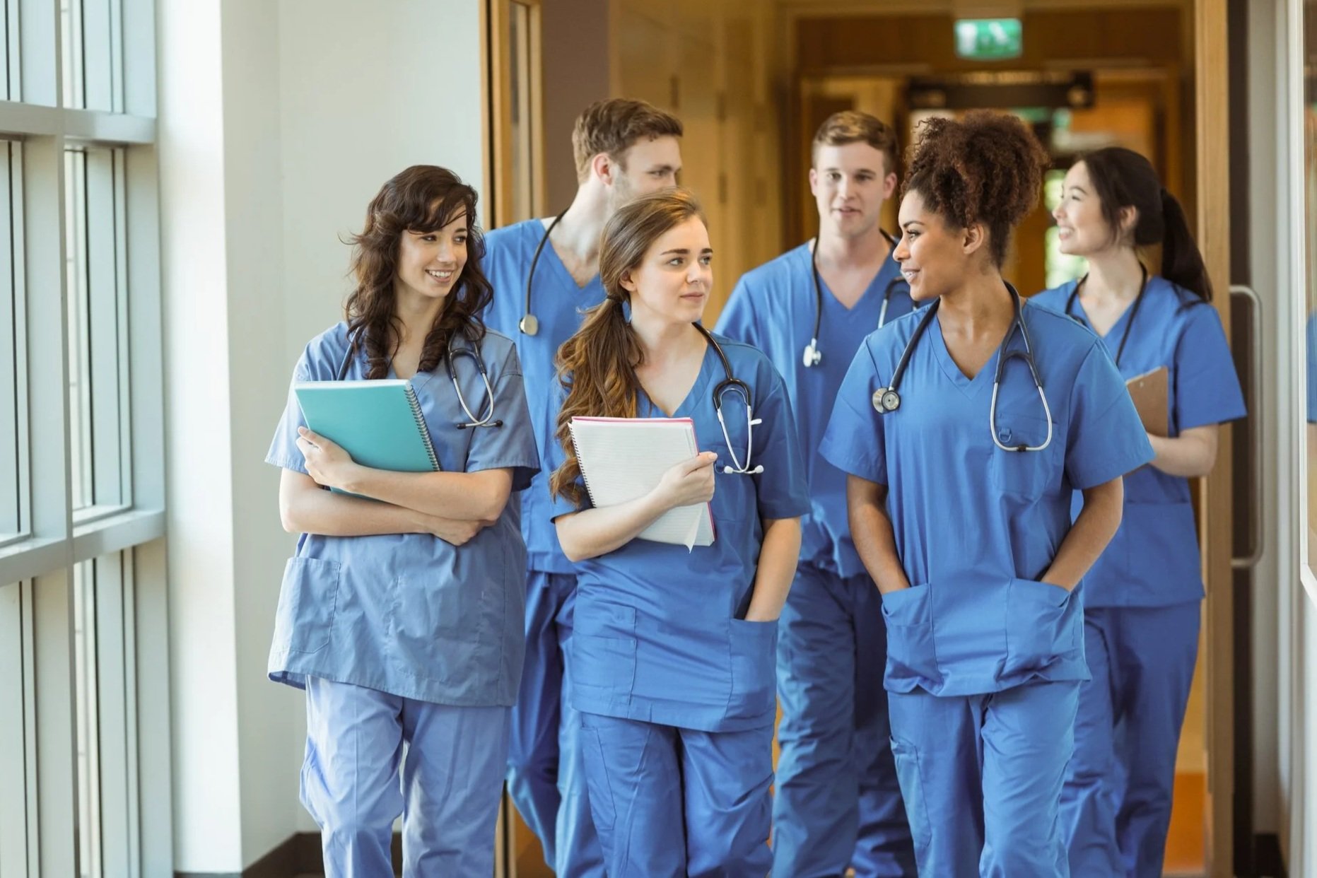 Group of six diverse healthcare professionals, dressed in blue scrubs, walking and talking in a hospital corridor.