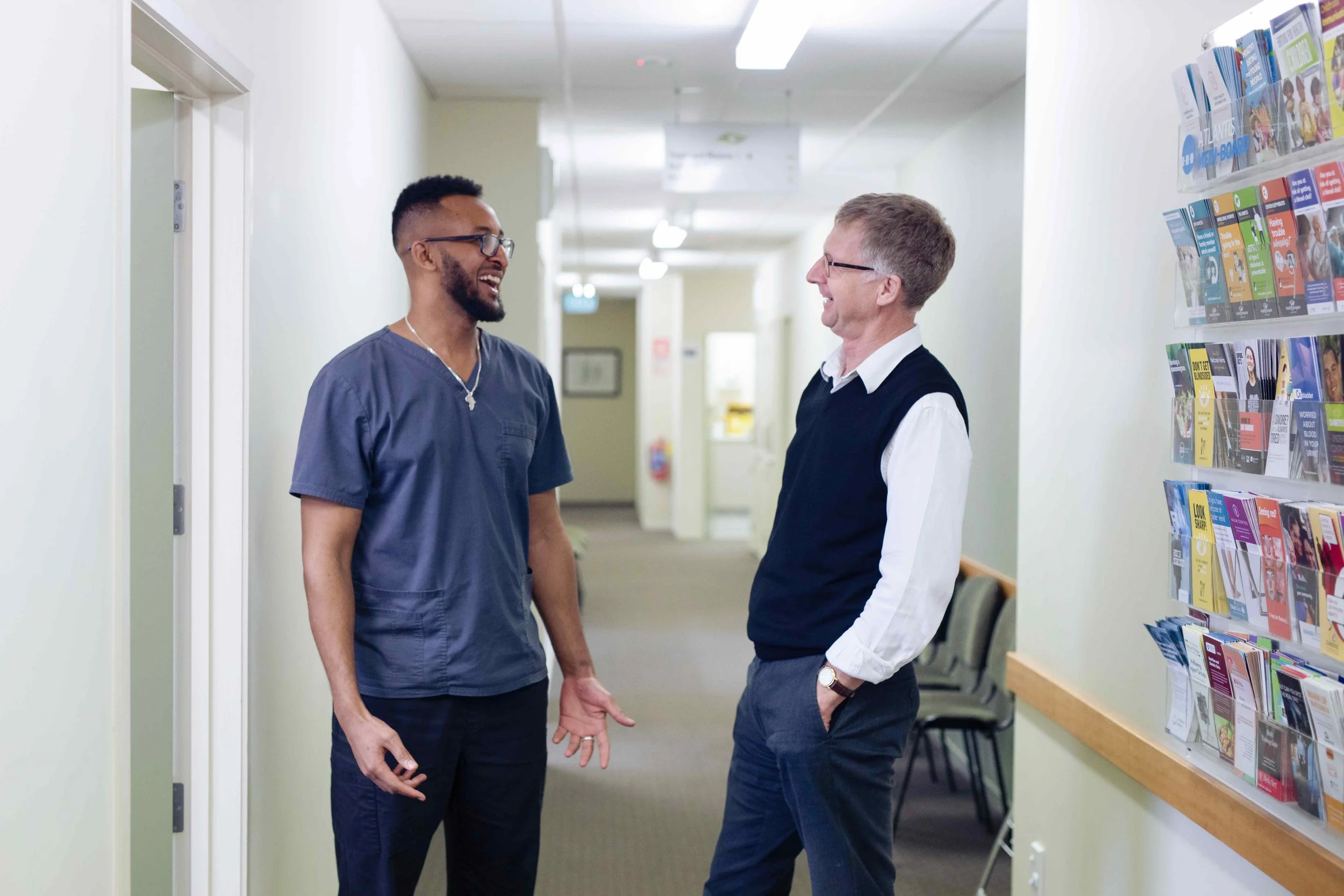 Two men are standing and talking in a hallway, smiling and making eye contact. One man is wearing scrubs and glasses, and the other is wearing a vest and white shirt. There is a bookshelf with pamphlets on the right side and chairs in the background.
