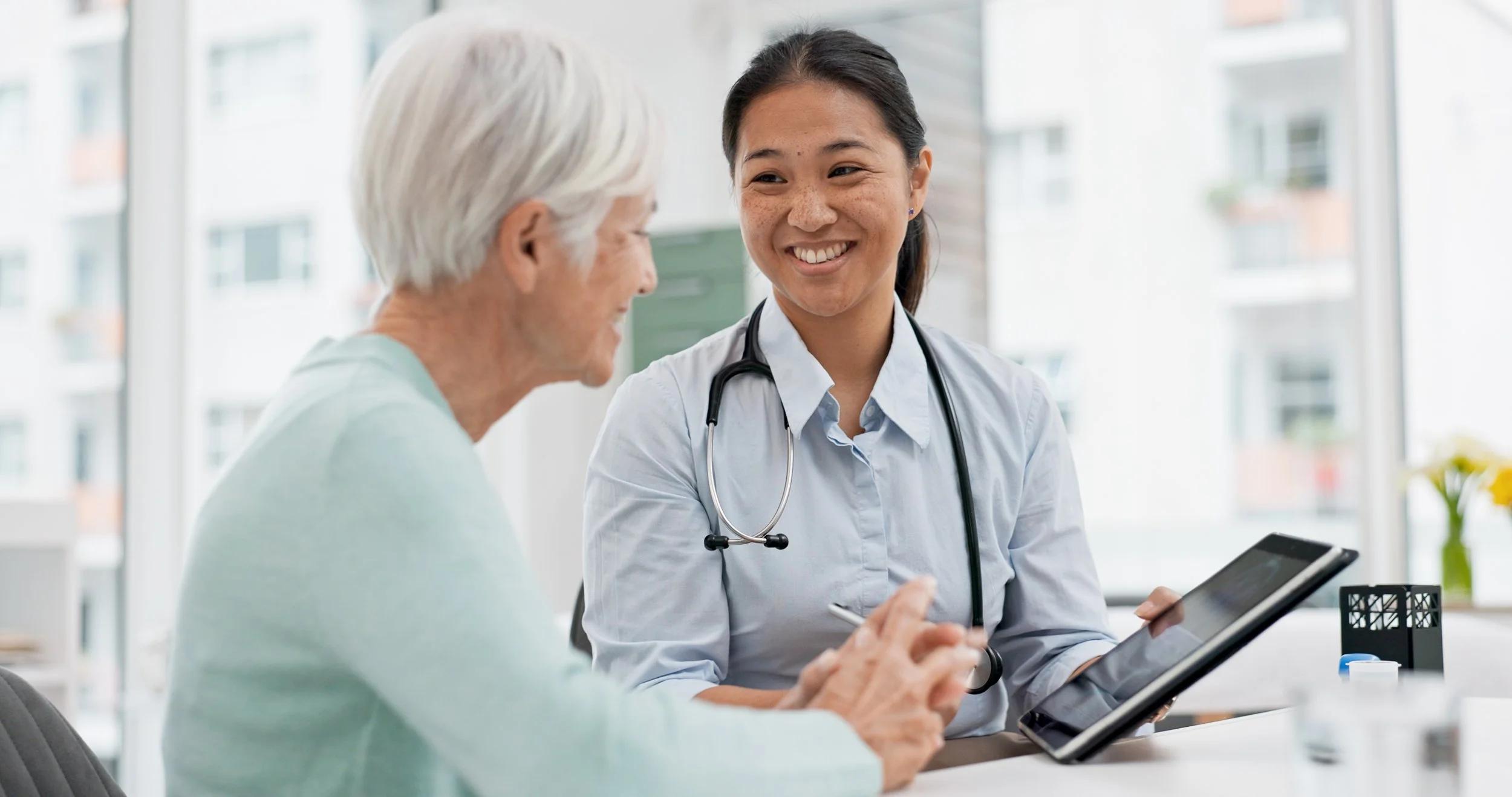 A doctor talking with an elderly patient in a medical office, both smiling, with the doctor holding a tablet.