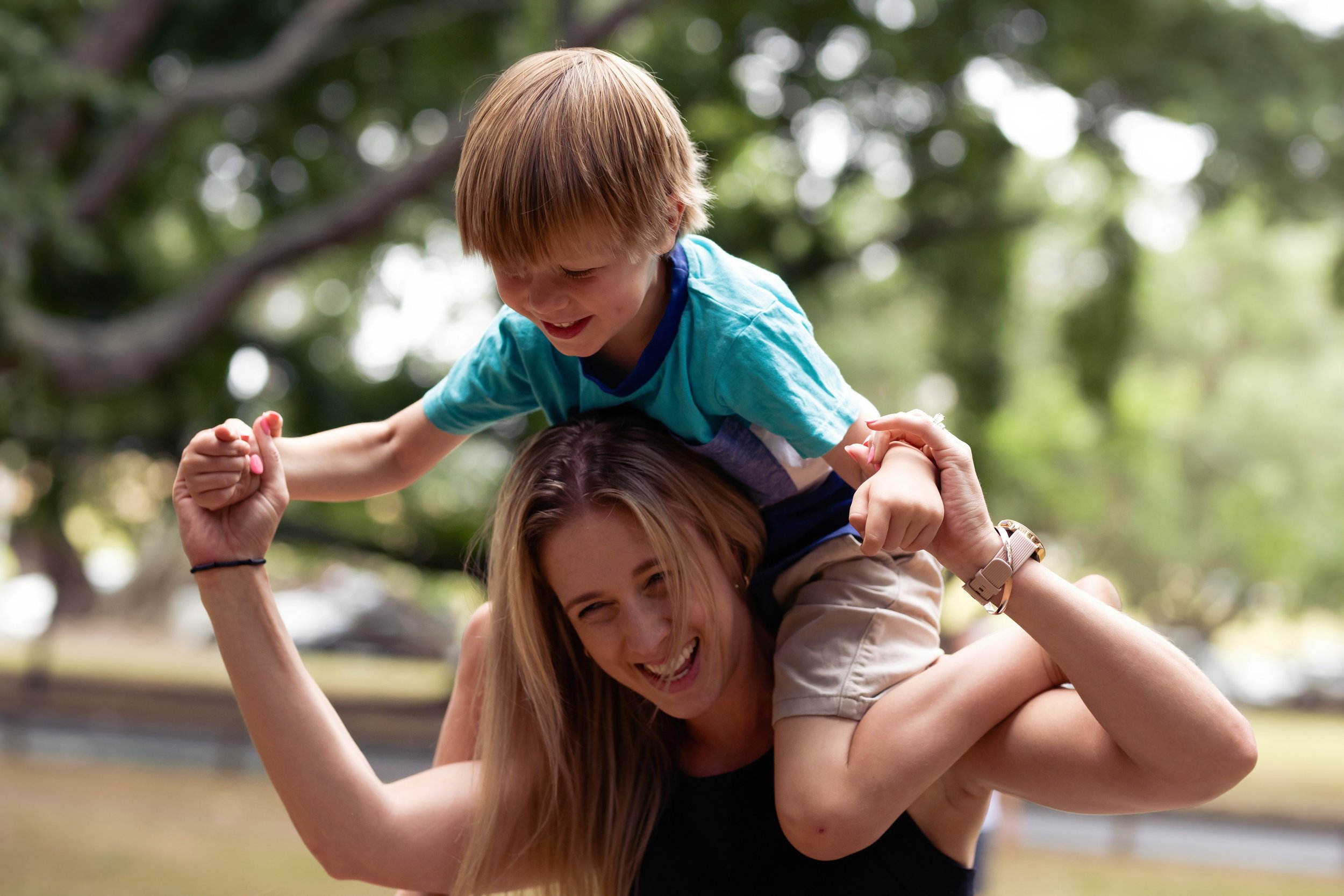 A woman with long hair smiling and holding a young boy on her shoulders outside in a park. The boy has blonde hair and is wearing a blue shirt, smiling and looking down. The background shows blurred green trees and sunlight.