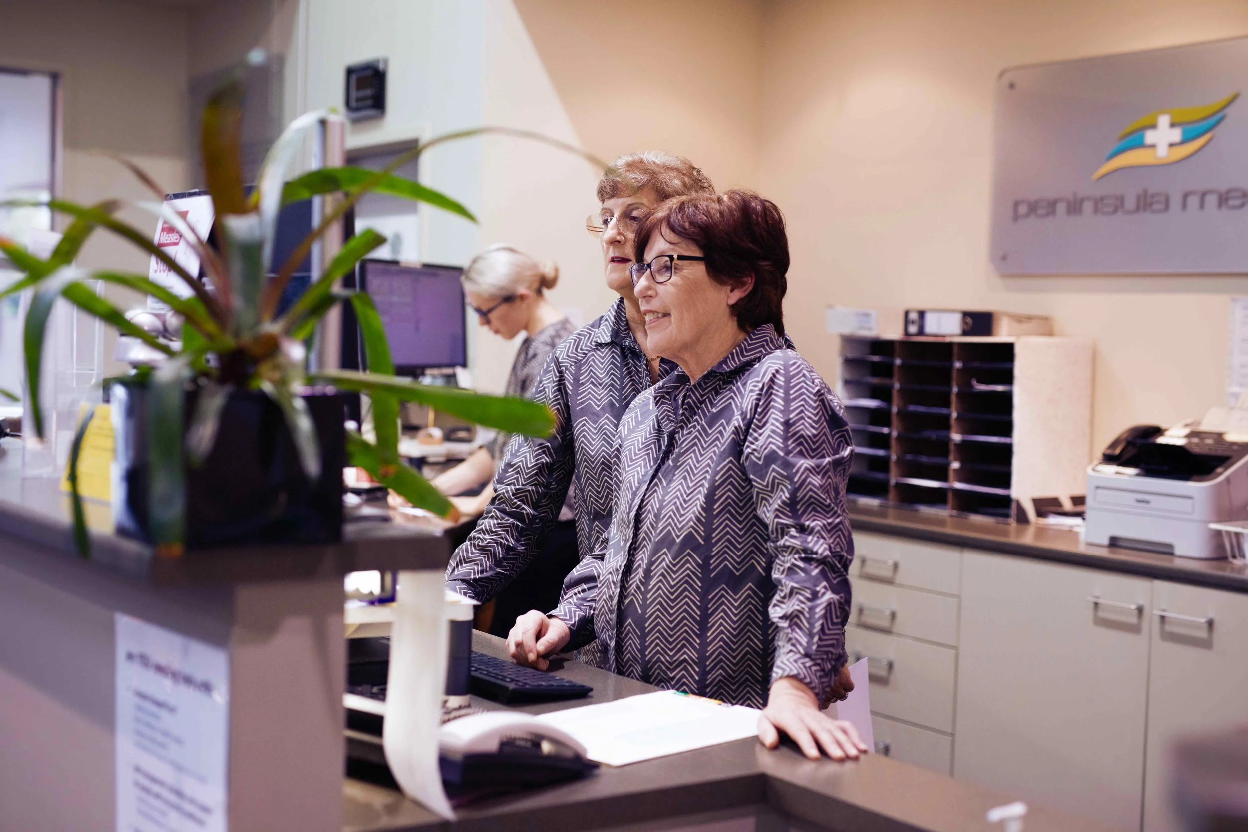 Two women, one with glasses and short dark hair, and the other with curly hair, working at a medical reception desk with computers and office supplies. A third woman with glasses is working in the background. There is a sign on the wall with a medical logo and the text "PENINSULA" and "MED".