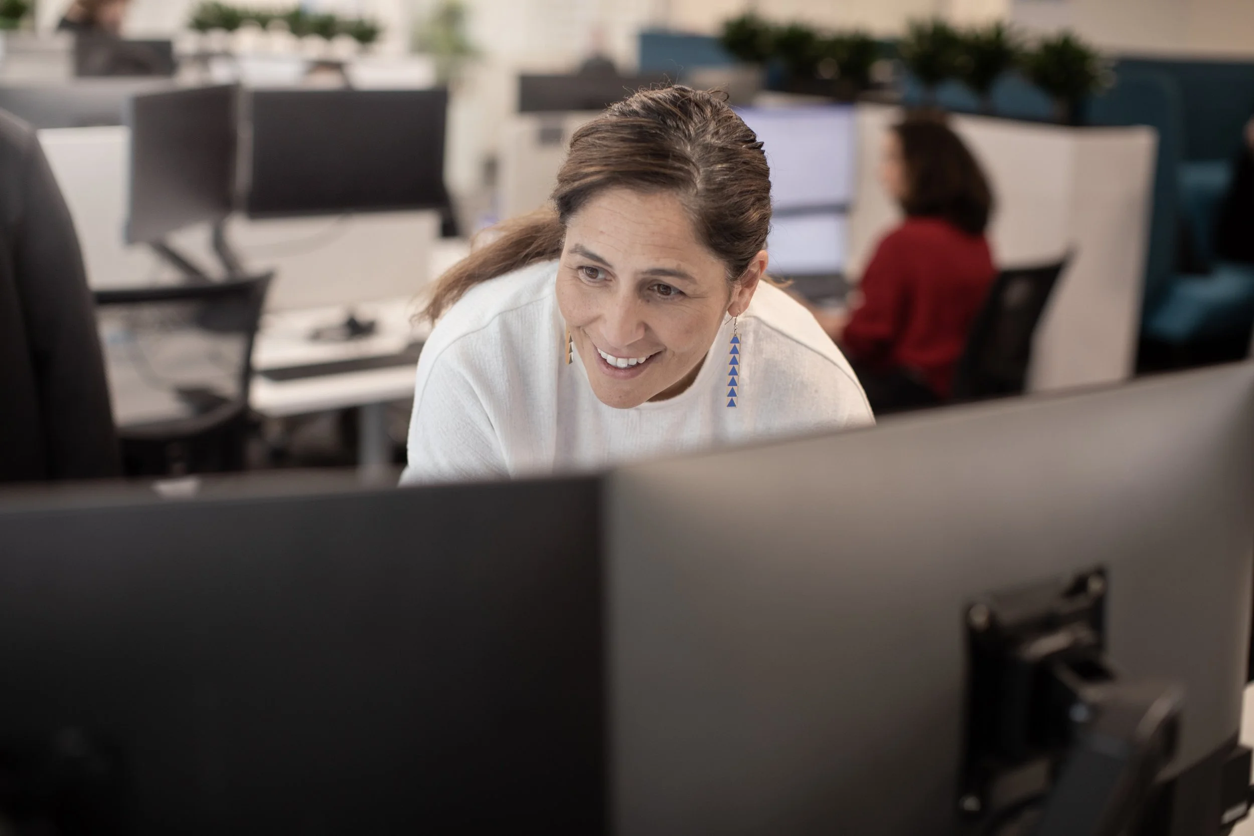 A woman with brown hair and earrings, smiling while working at her computer in an office environment, with another person blurred in the background.