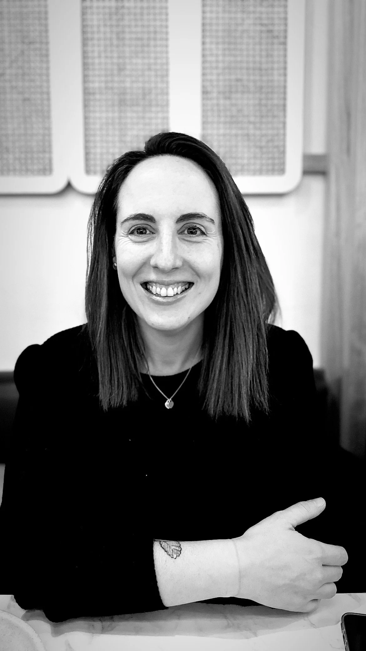 Black and white photo of a woman with shoulder-length hair smiling, wearing a black top and a necklace, with her arm resting on a table showing a small tattoo of a leaf on her wrist, and a smartphone on the table.