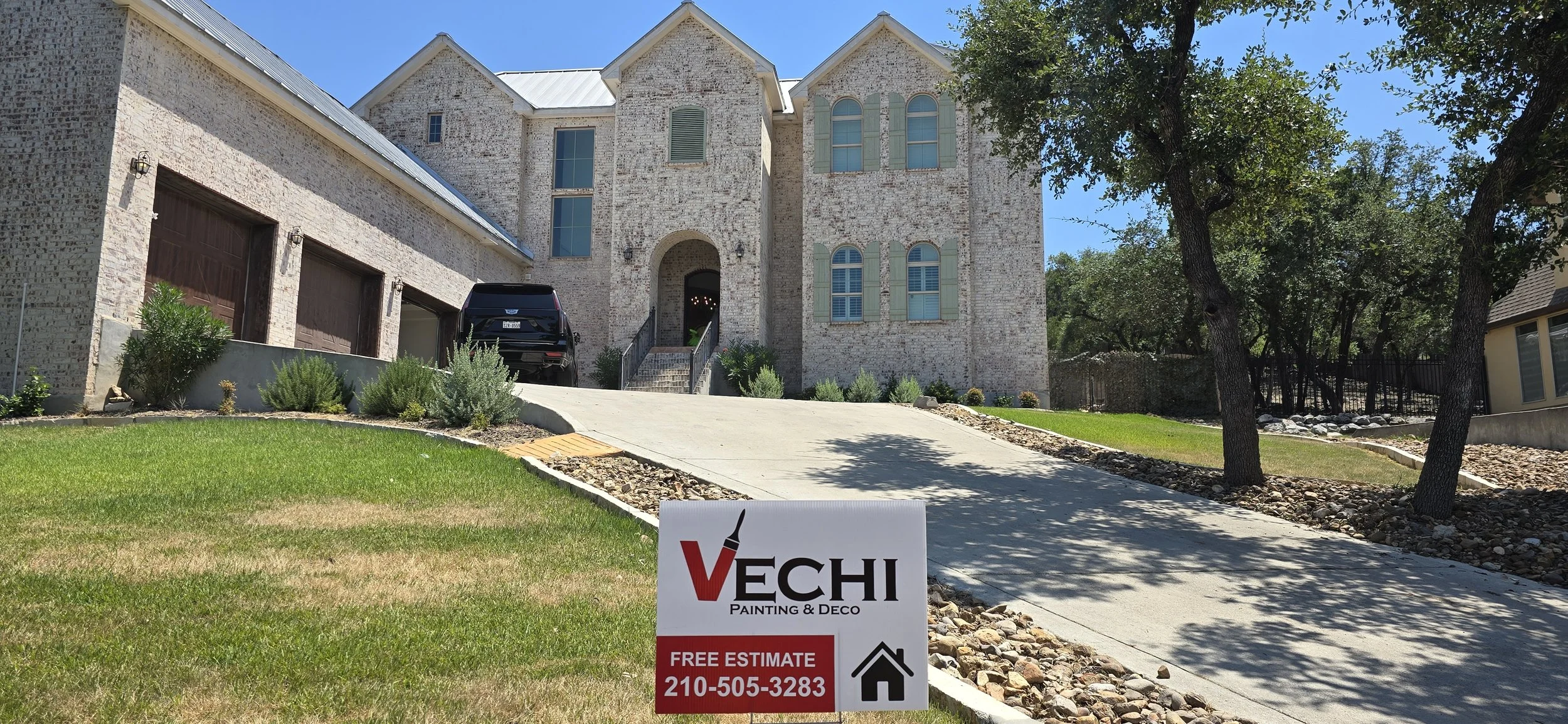 A large brick house with a driveway and a black car parked near the garage. An advertisement sign in the foreground for Vechi Painting & Deco with a phone number and a house icon.