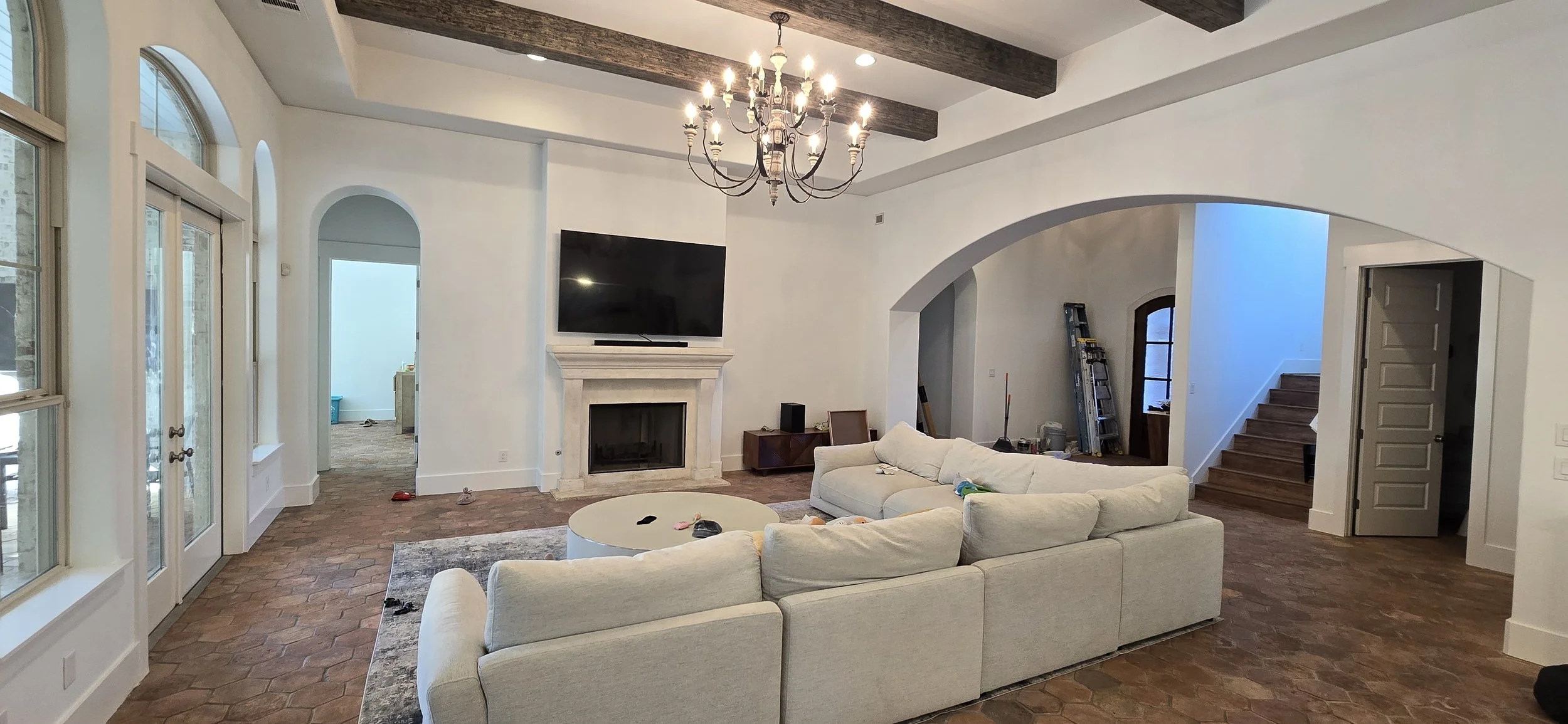Modern living room with white walls, a chandelier, a large off-white sectional sofa, a round coffee table, a fireplace with a TV mounted above it, all on a brick floor, with stairs leading up on the right.