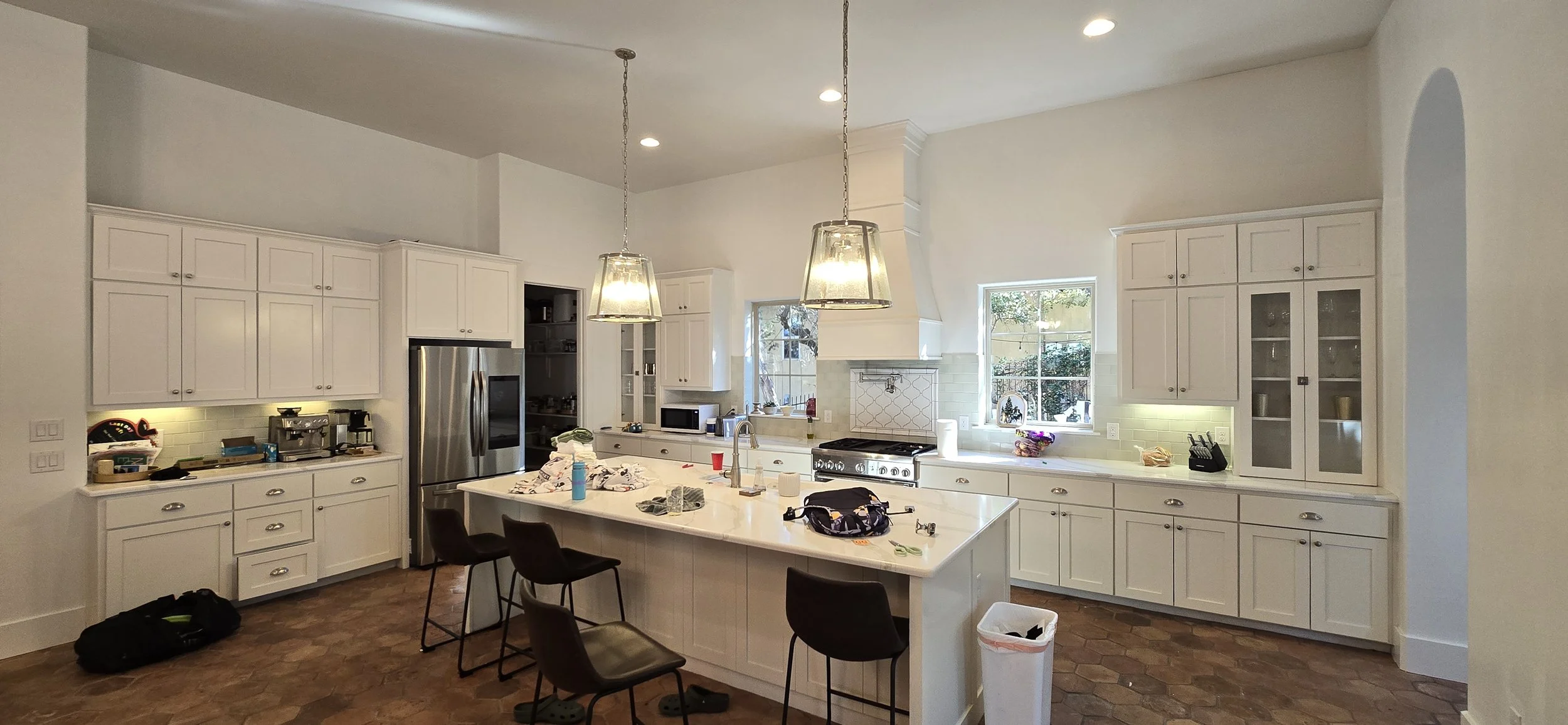 Modern kitchen with white cabinets, stainless steel refrigerator, island with black chairs, and two hanging pendant lights, featuring various appliances and clutter on countertops.