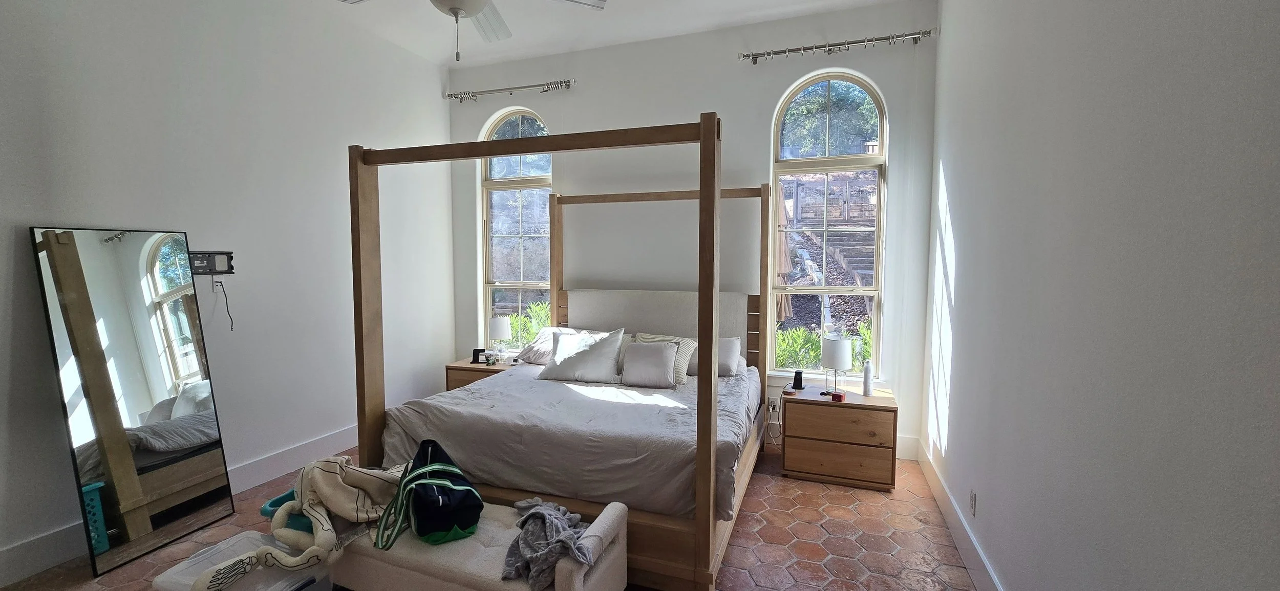 Bedroom with a wooden four-poster bed, two tall arched windows, and a large mirror leaning against the wall reflected in the window, with a ceiling fan and terracotta tile floor.
