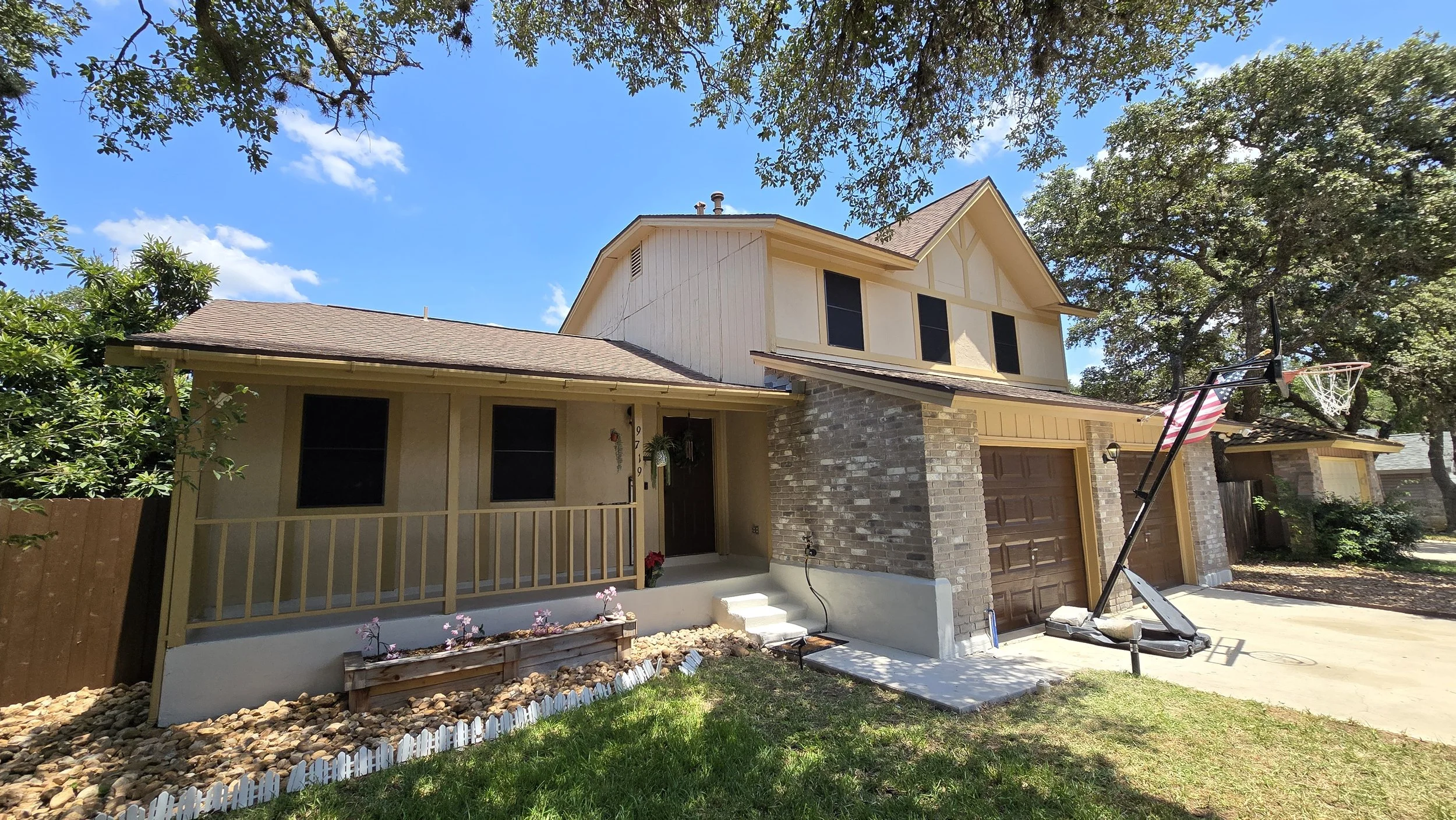 A two-story house with a brick and yellow siding exterior, a brown garage door, and a small front porch with a railing. There is a portable basketball hoop set up near the driveway, and an American flag attached to it. The house is surrounded by trees and a well-maintained lawn.