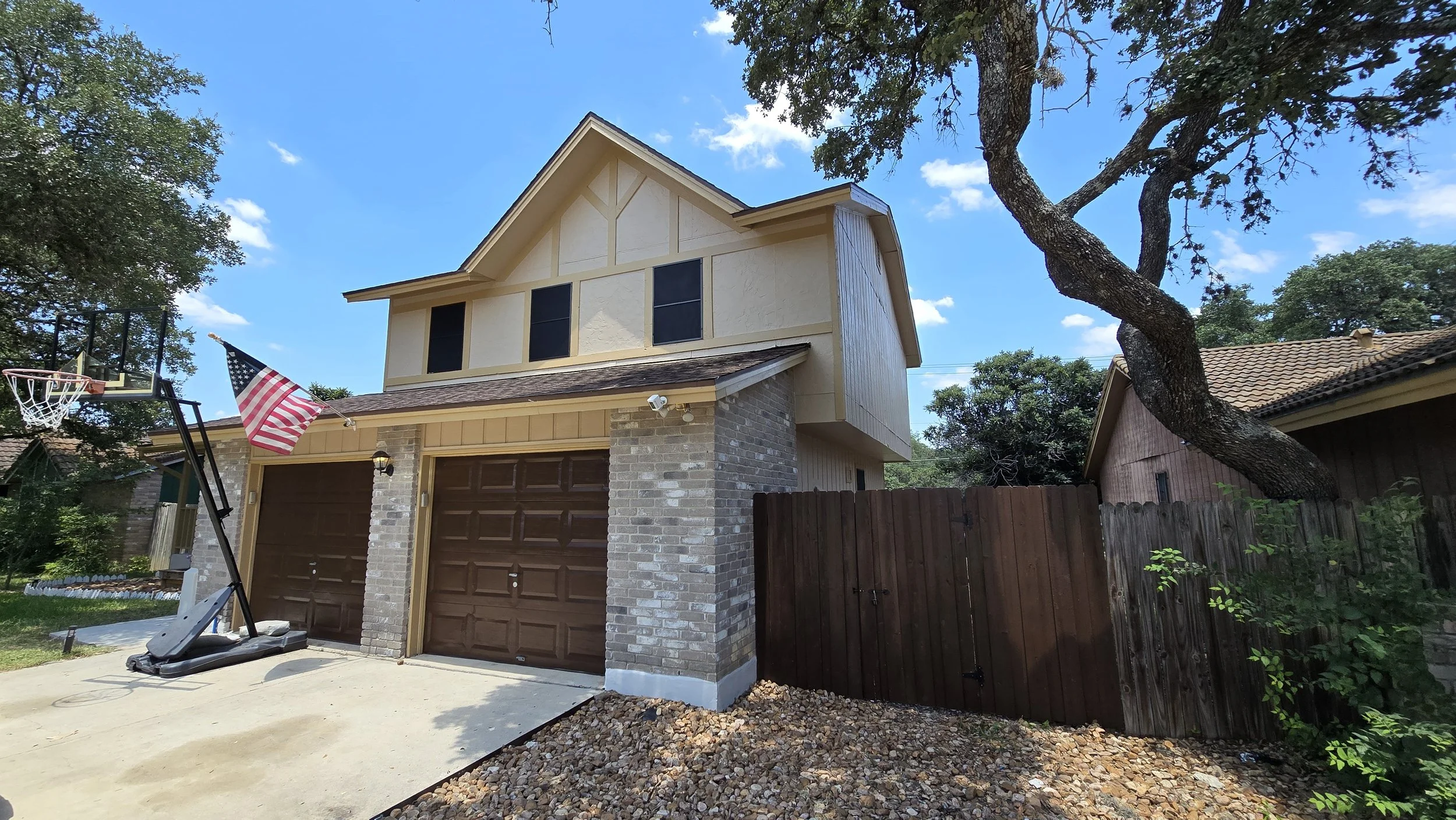 Two-story house with brick and cream siding, brown garage doors, American flag on a pole, and an outdoor basketball hoop with a backboard and a portable base in the front yard, with a large tree to the right, under a blue sky with a few clouds.