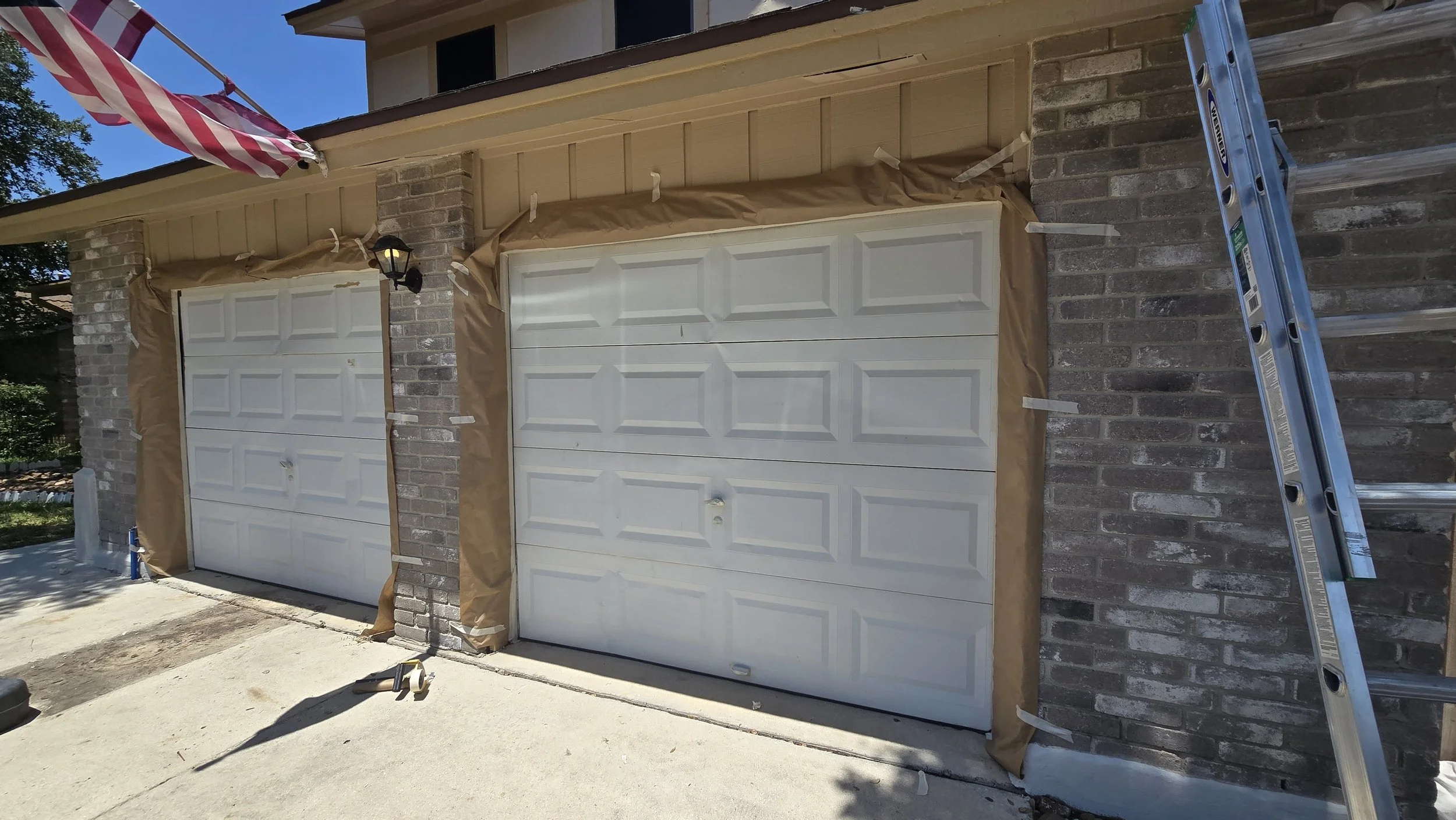 Two garage doors under maintenance, with brown paper and tape around the edges, a ladder on the right side, and a shadow of a hammer on the ground, in front of a brick house with an American flag.