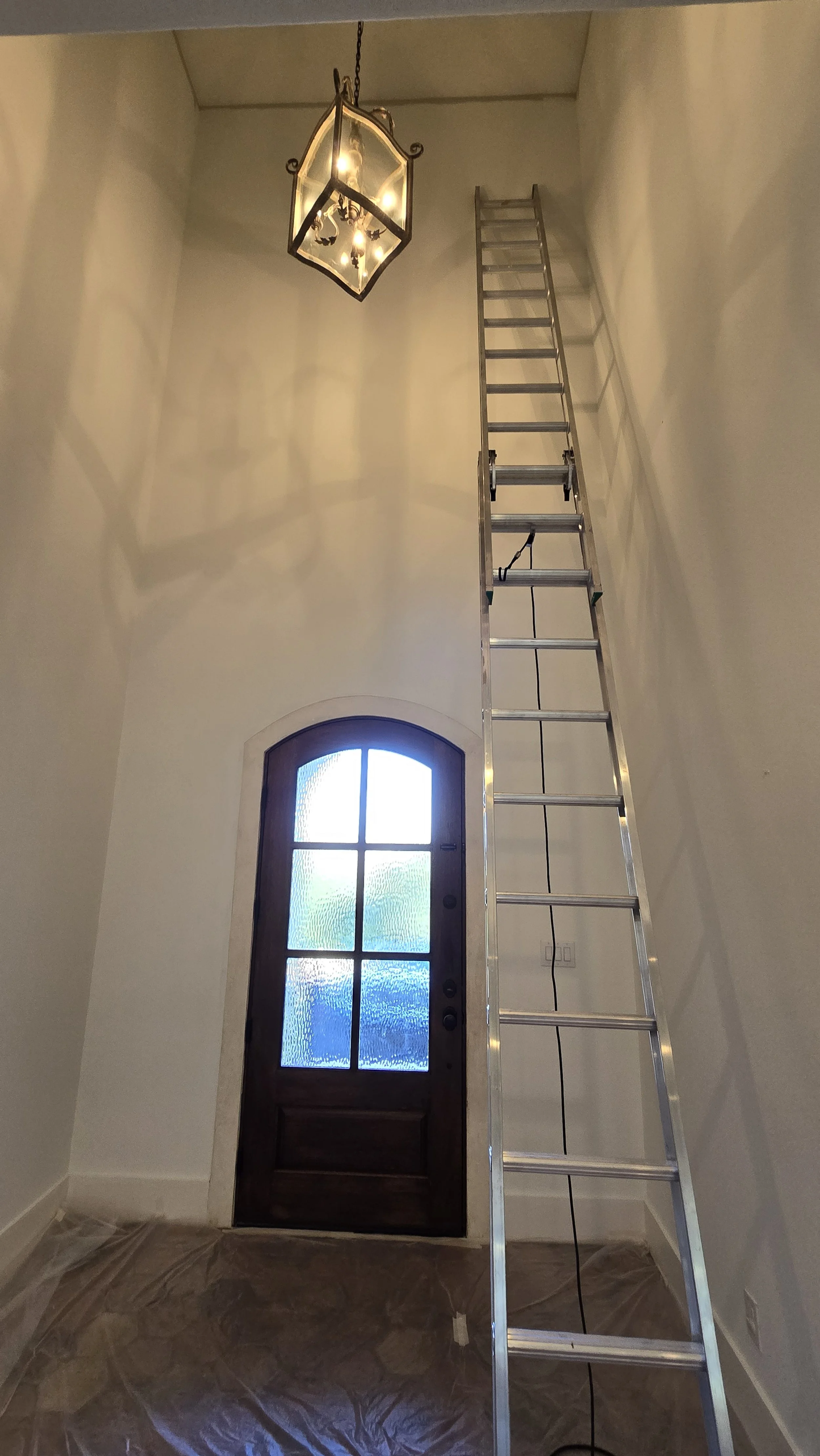 Interior view of a foyer with a dark wooden front door with frosted glass panels, a ladder reaching the ceiling, a chandelier hanging from the ceiling, and protective covering on the floor.