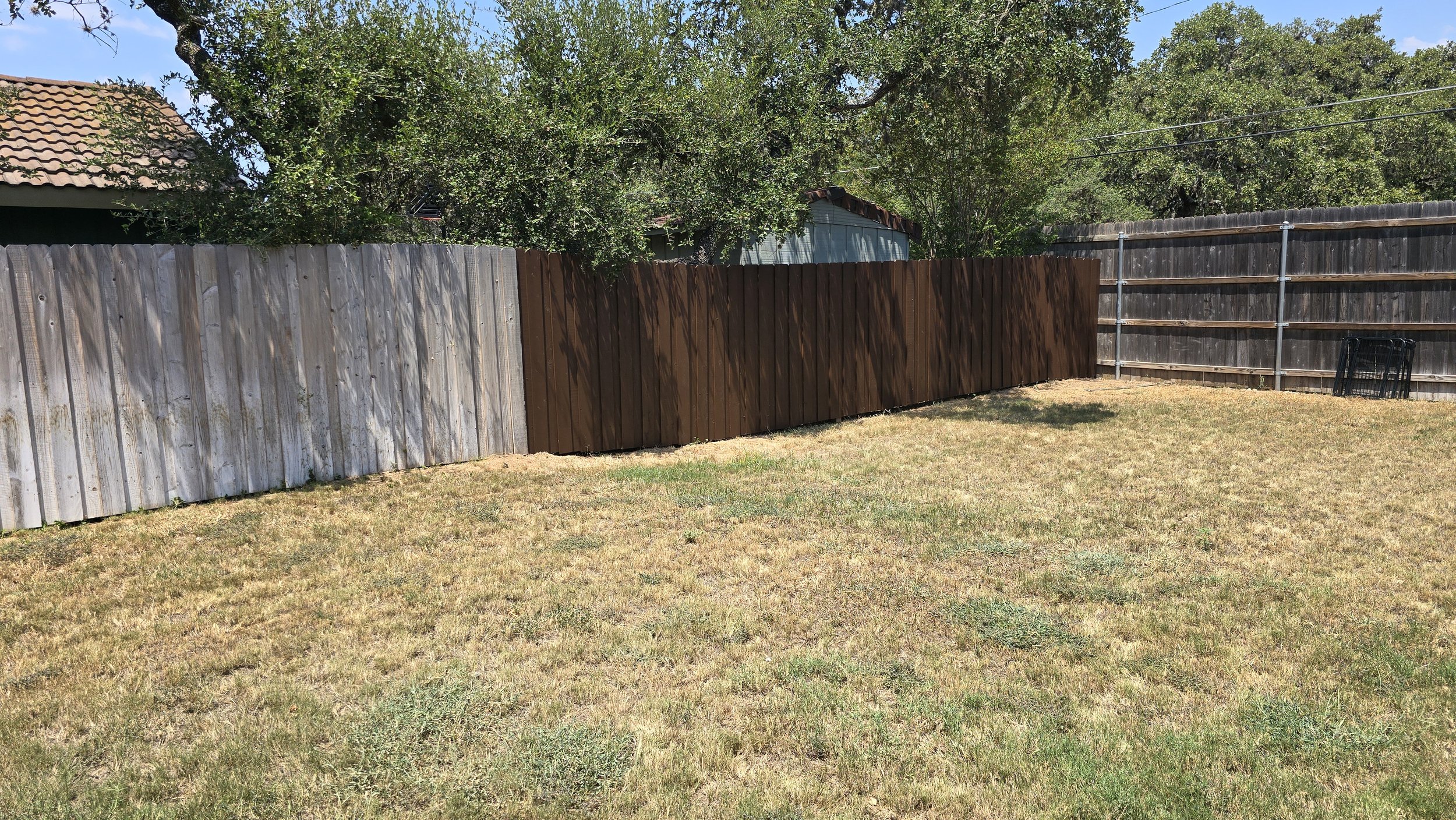 Backyard with a grassy lawn, a gray wooden fence on the left side, and a brown wooden fence in the middle, with trees and a shed visible behind the fences.