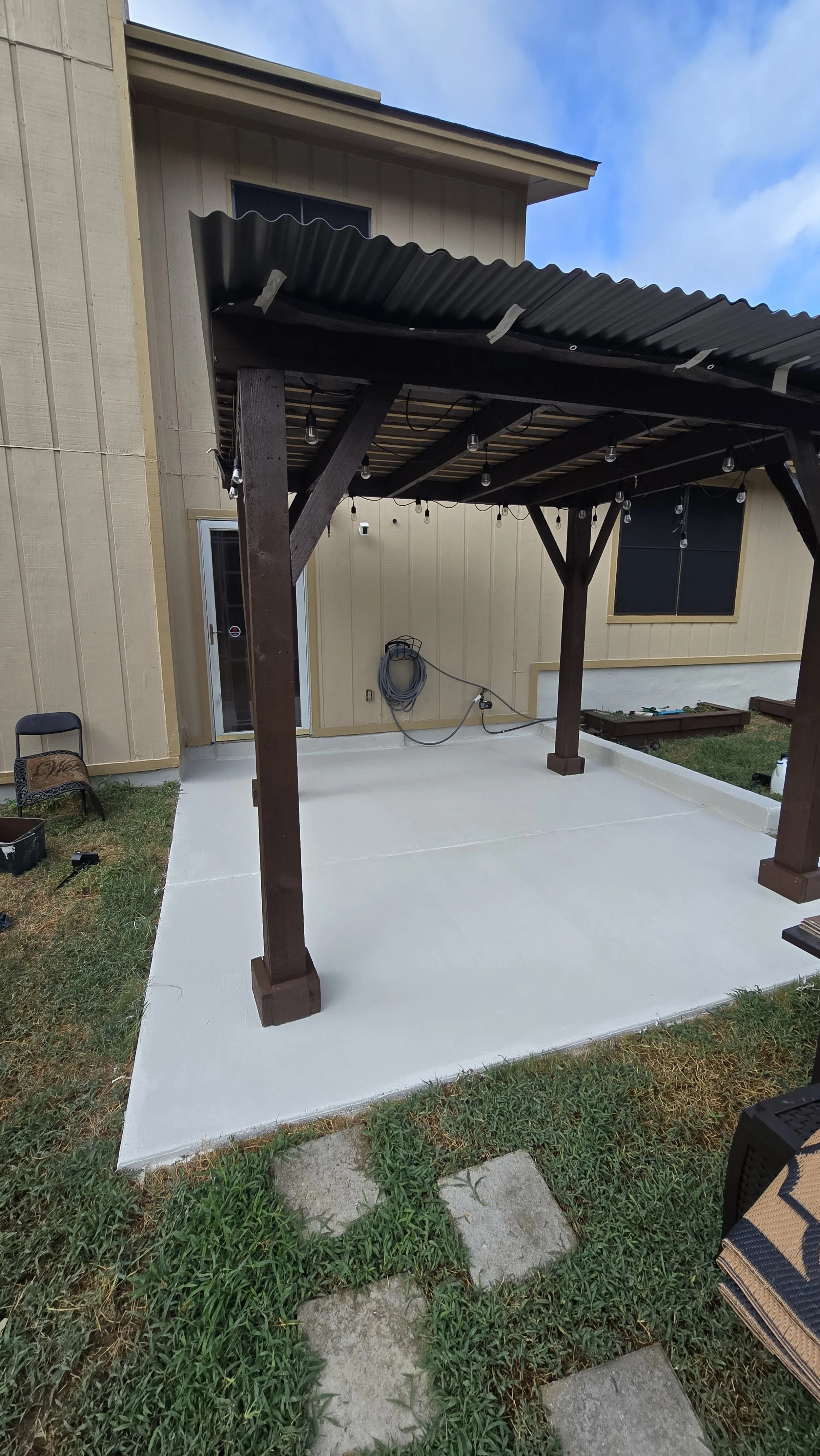 Backyard patio with a newly finished concrete floor, a wooden pergola with string lights, a hose reel on the wall, and a sliding glass door leading into the house.