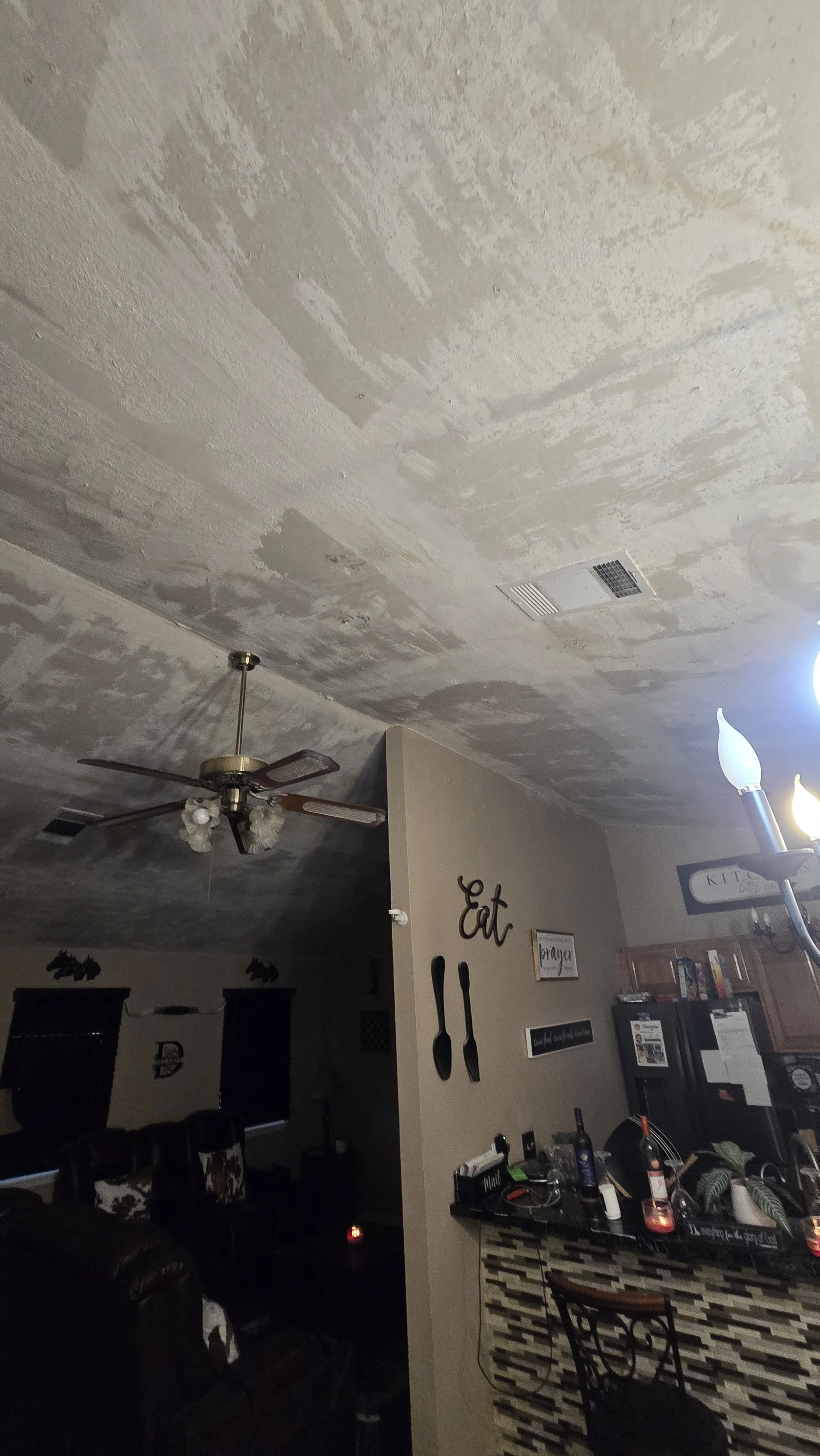 Unfinished ceiling with patches of drywall mud; ceiling fan and kitchen chandelier visible; living room and kitchen area with furniture and decorations in the background.