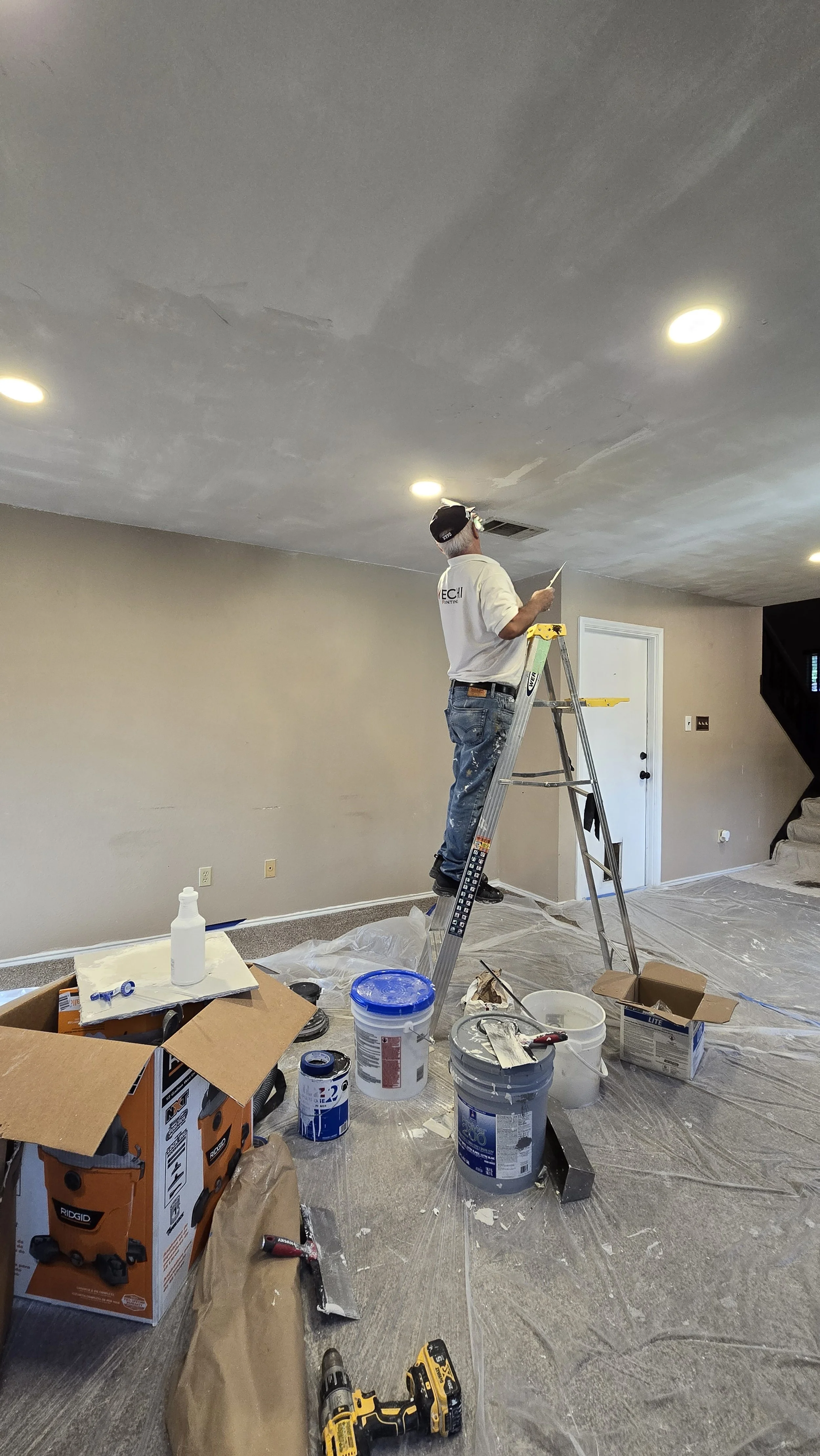 A man on a ladder painting a ceiling with a paintbrush in a room under renovation, surrounded by paint buckets, tools, and plastic covering the floor.