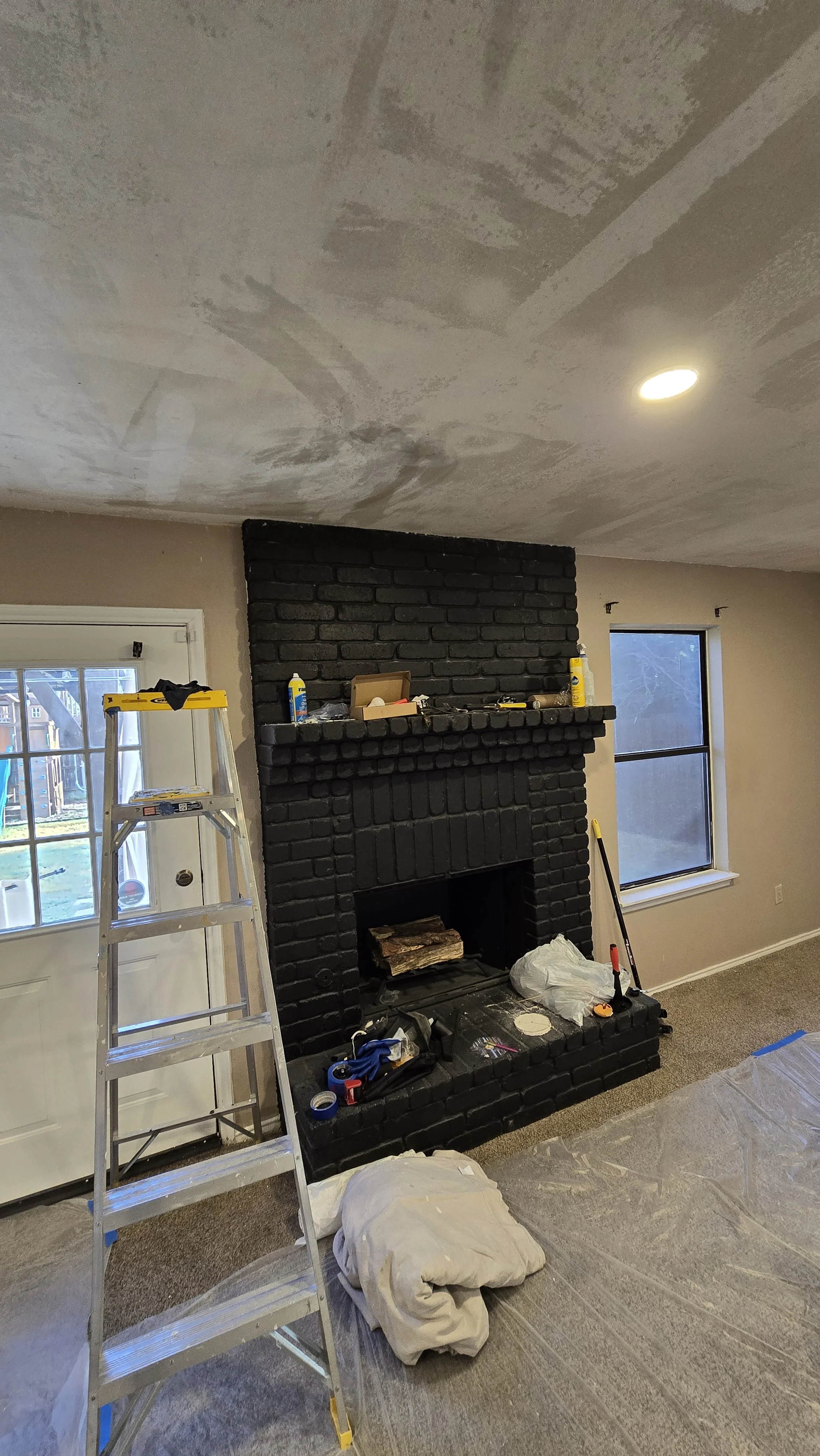 Living room under renovation with a black brick fireplace, ladder, and tools. The ceiling appears to be freshly patched or painted.