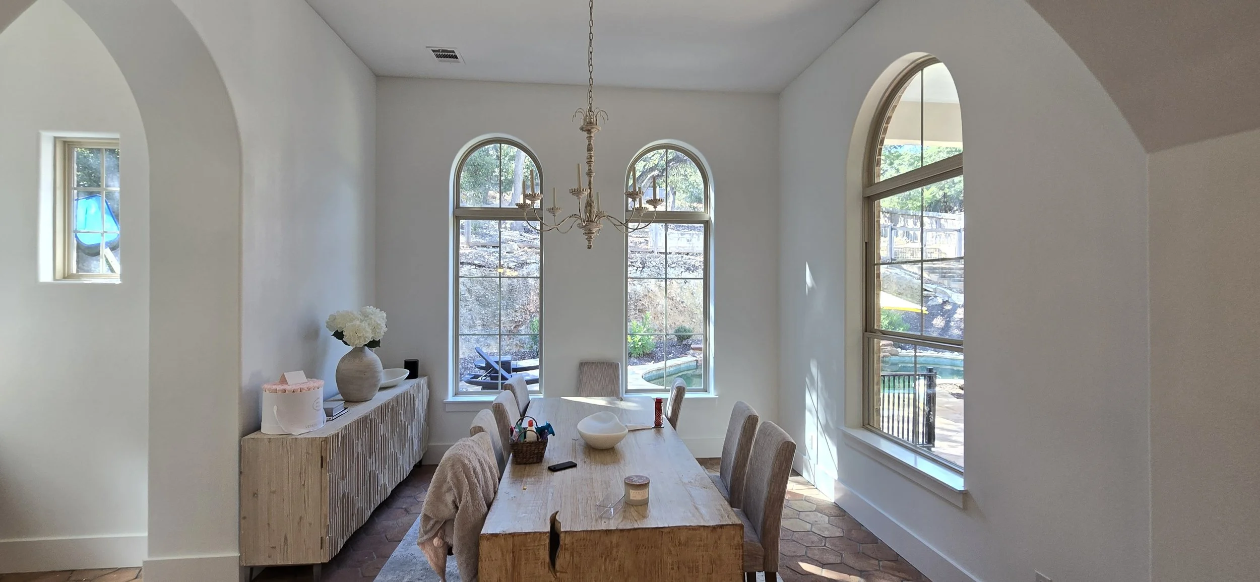 Dining room with a large wooden table, six upholstered chairs, a chandelier, three arched windows, a sideboard with a vase of flowers, and a view of the backyard.