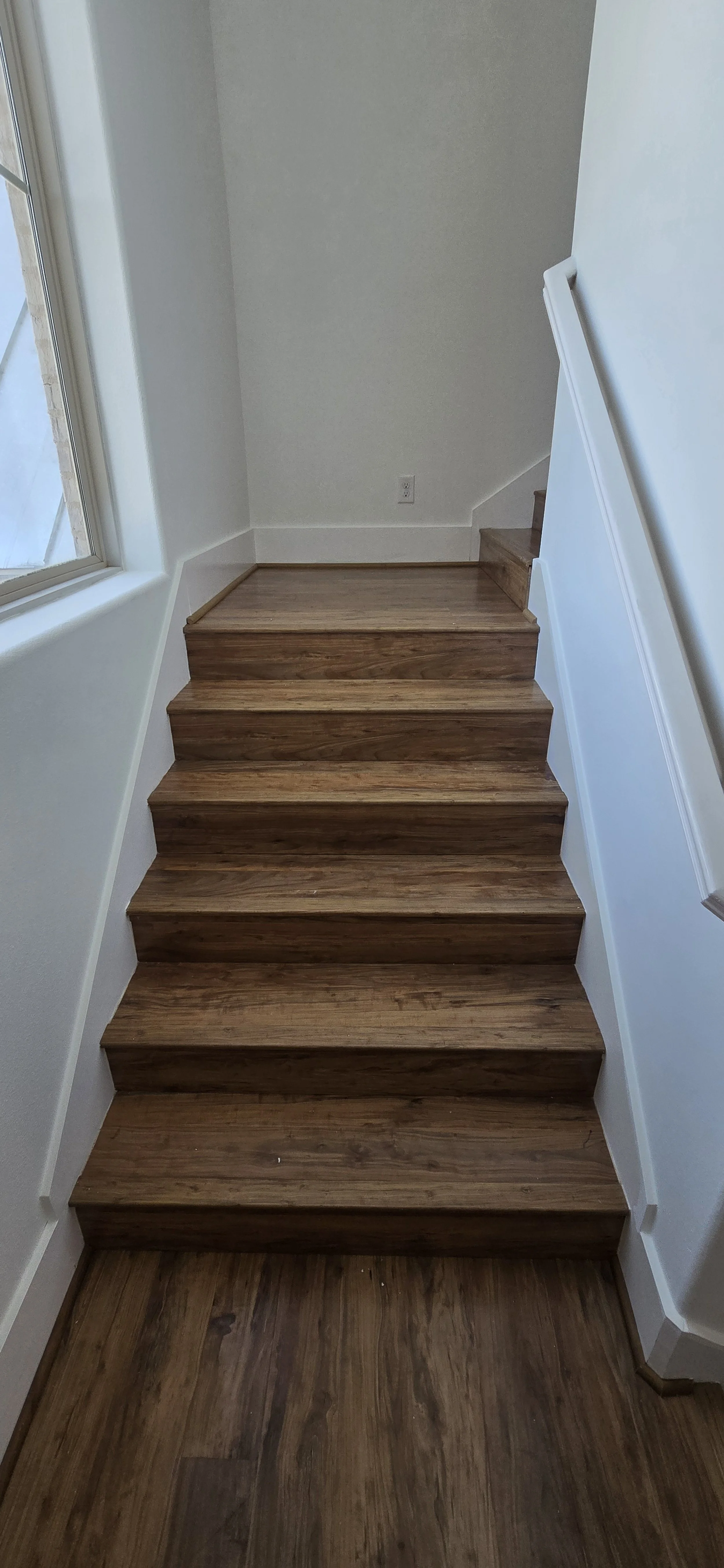 Image of a staircase with wooden steps and a white wall on the right side, next to a window on the left, in a well-lit area.