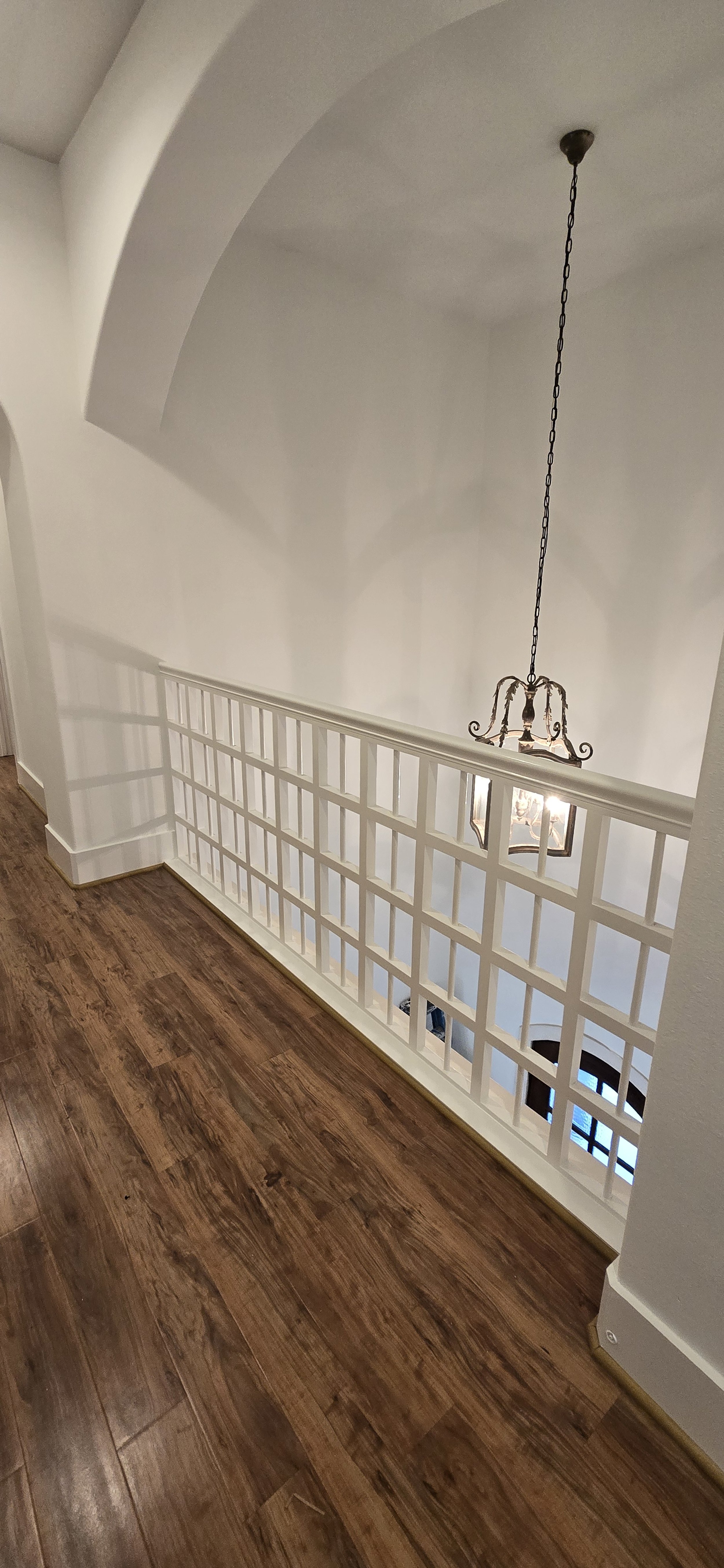 A view of an upstairs hallway with wooden flooring, a white railing with multiple square cutouts, a large hanging chandelier, and an arched ceiling.