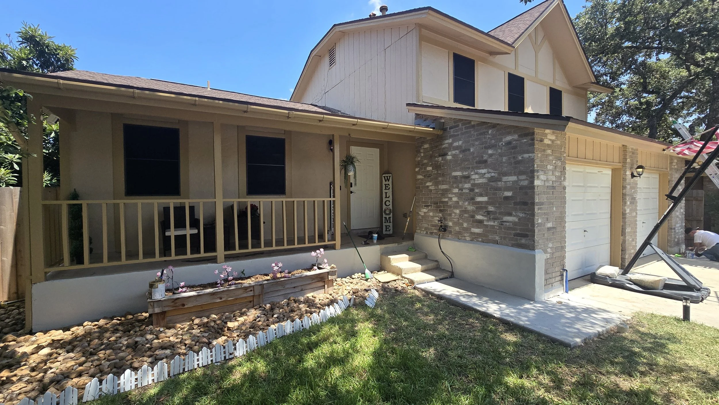 Two-story house under construction with an unfinished porch and landscaping in progress; workers are seen on the right working near the driveway.