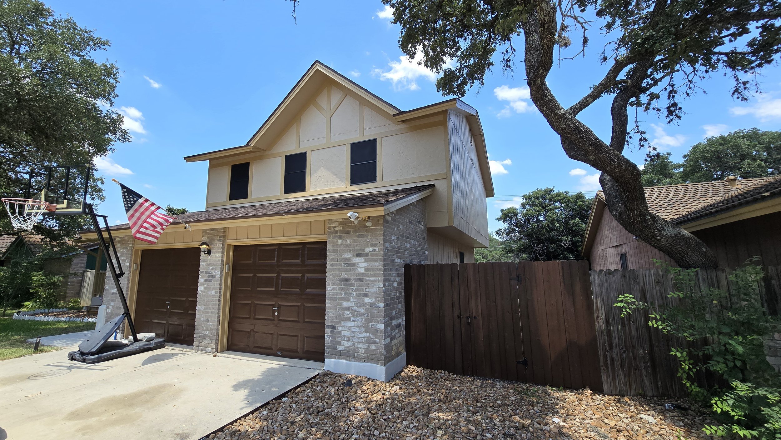 The front of a two-story house with a double garage, brick walls, and beige siding. An American flag hangs on a pole attached to the house, and there is a basketball hoop and stand in the driveway. A large tree with a twisted trunk leans over the house, and the sky is blue with scattered clouds.