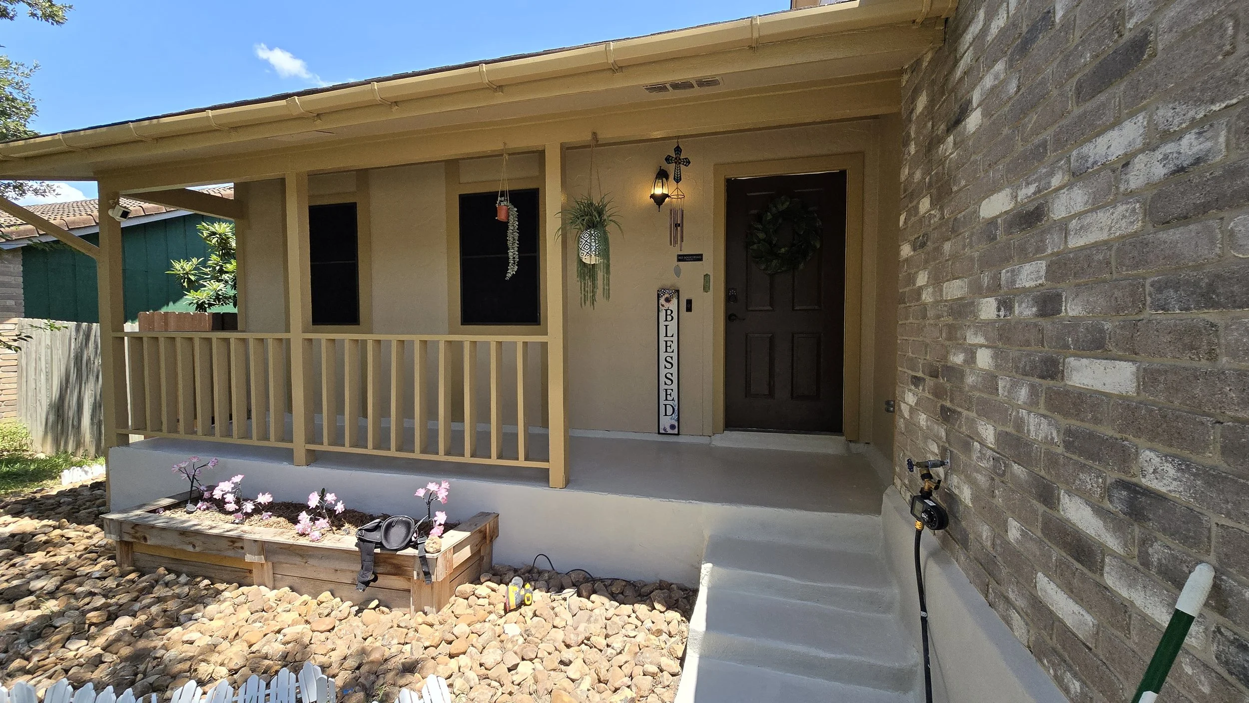 Front porch of a house with a dark wooden door, a wreath, hanging plants, and decorative items. A sign reading 'BLESSED' is next to the door, and there are steps leading to the porch. The yard has rocks and a wooden planter with pink flowers.