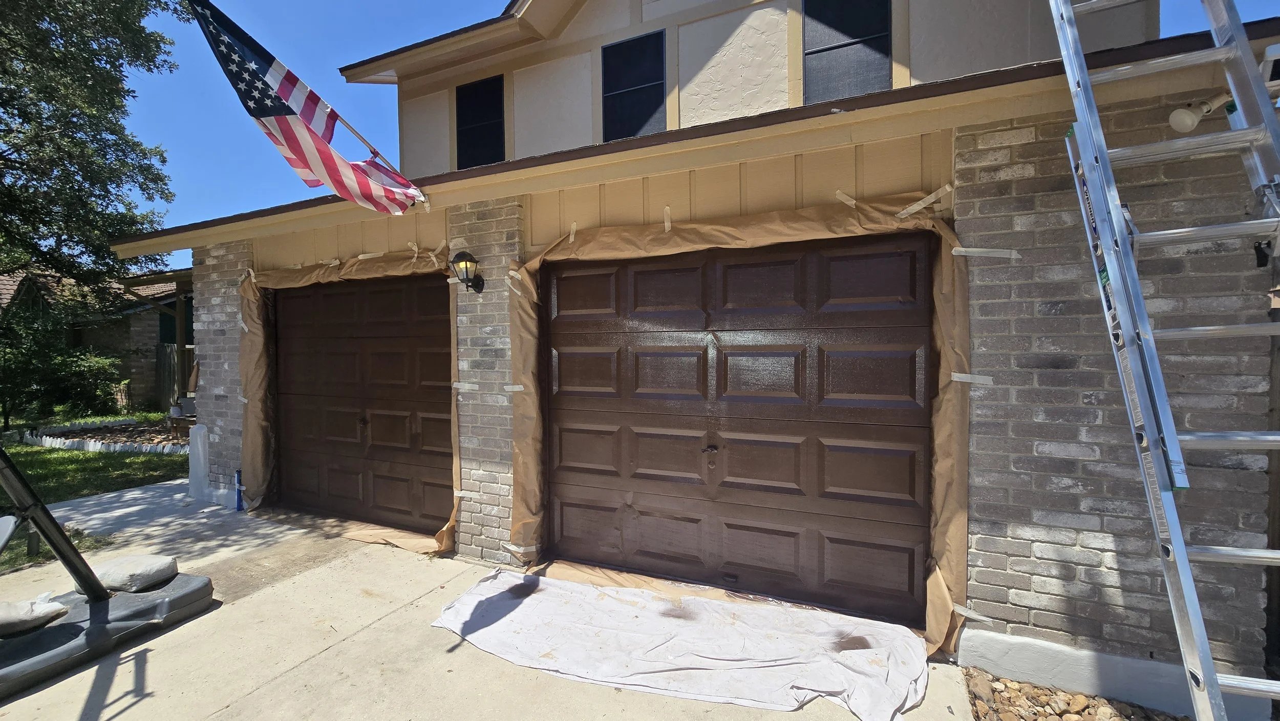 Two garage doors with brown wood-like finish, framed with paper tape for painting or staining, on a house with brick accents and beige siding. An American flag is mounted above, and a ladder is leaning against the house on the right.